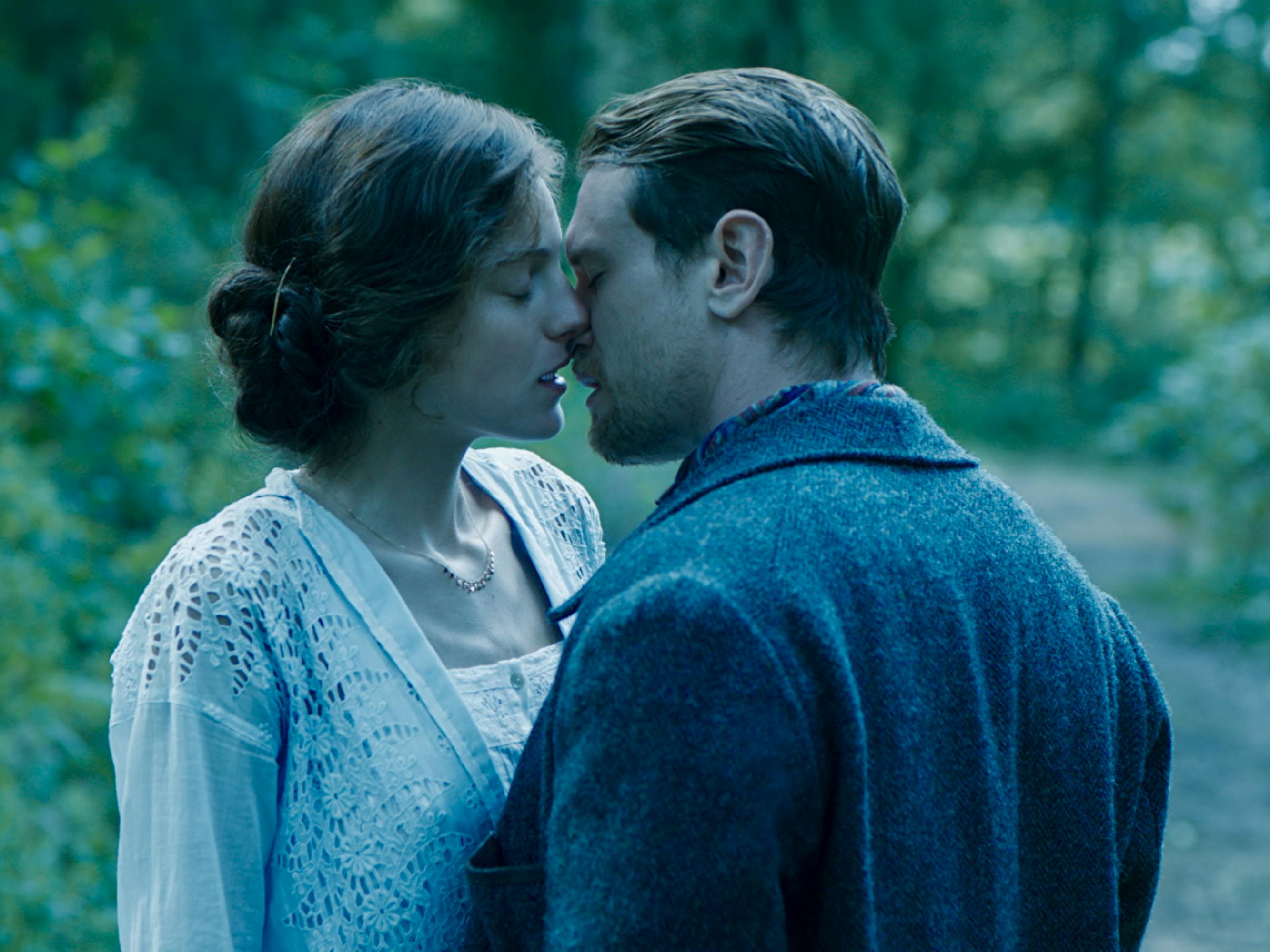 Lady Constance Chatterley (Emma) and Oliver Mellors (Jack O'Connell) embrace on a path shrouded with greenery.