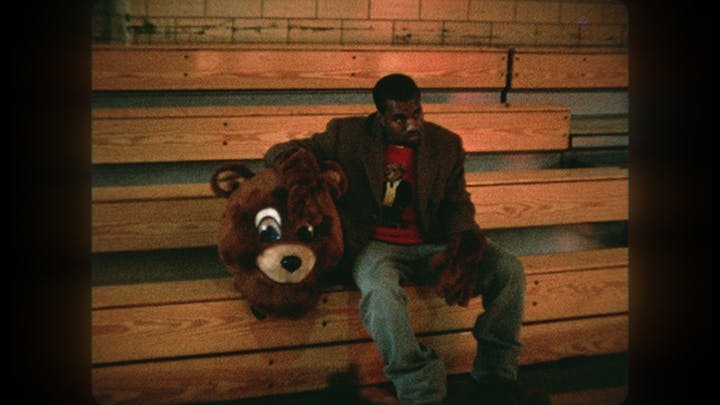 Kanye West wears jeans and a brown jacket and sits on wooden bleachers next to a massive stuffed bears head.