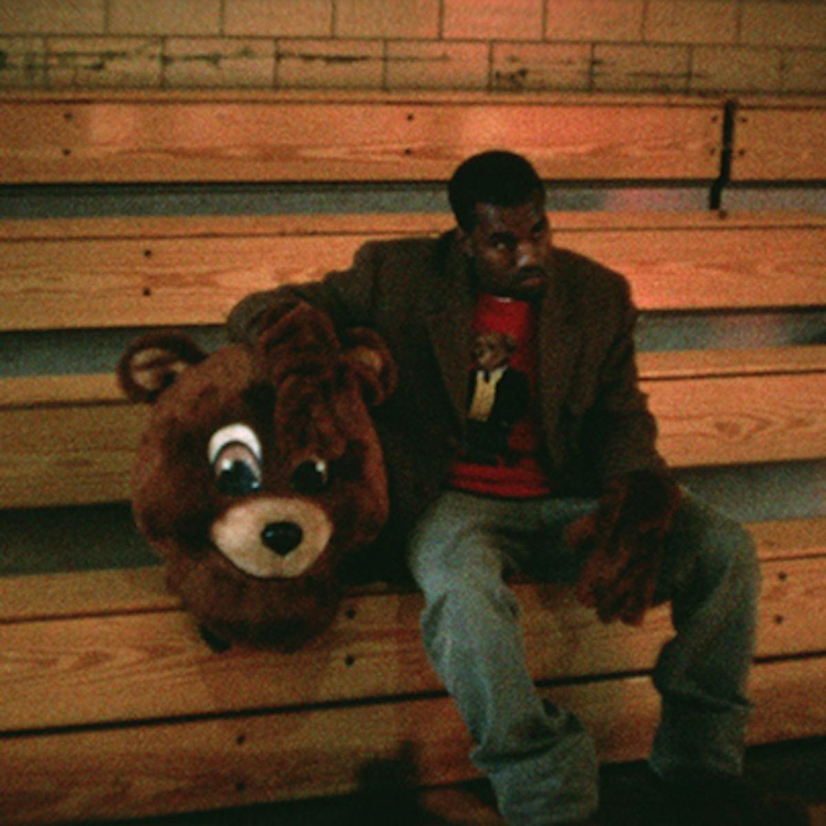 Kanye West wears jeans and a brown jacket and sits on wooden bleachers next to a massive stuffed bears head.