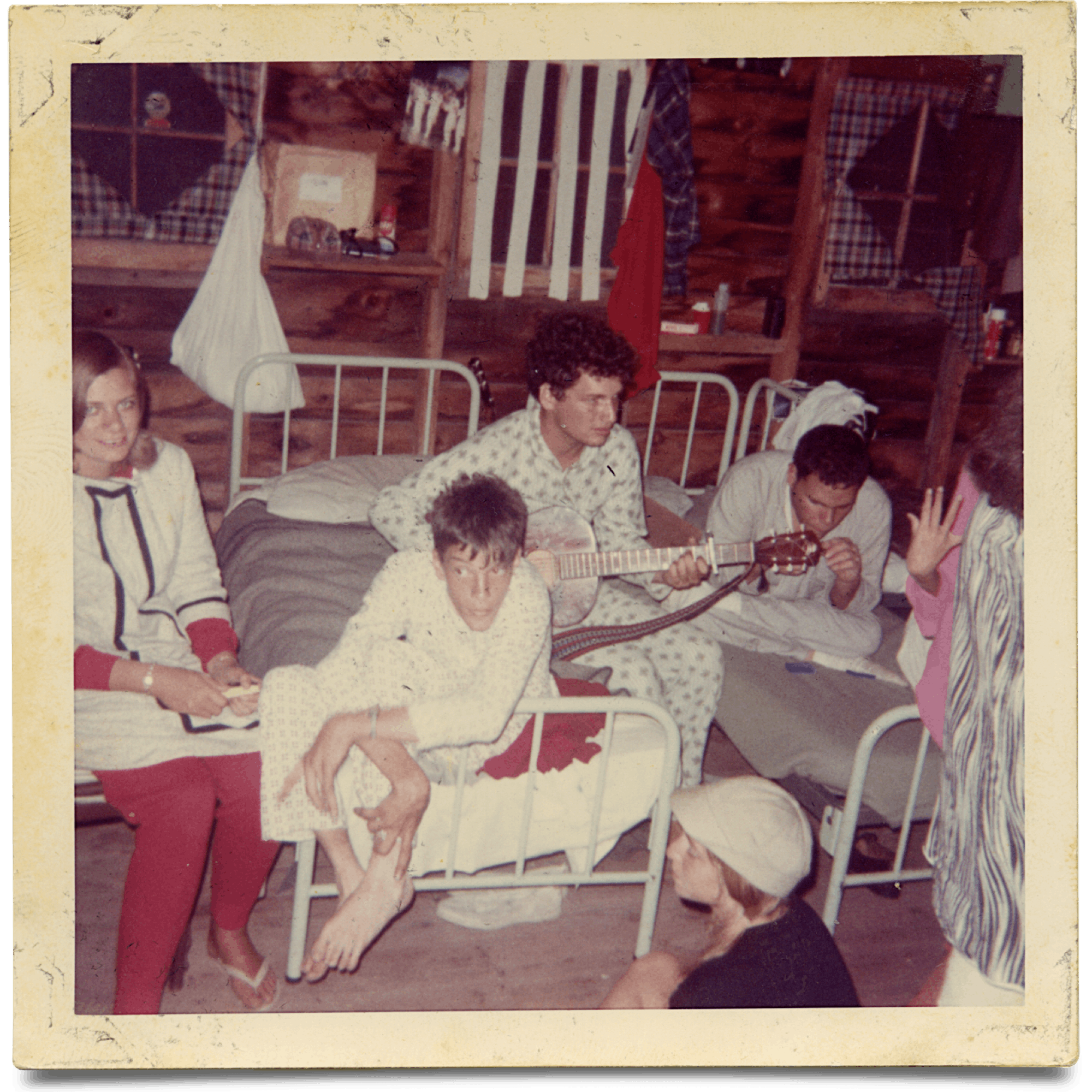 In an old photograph, teens are pictured perched around a cabin in their pajamas. One camper plays guitar.