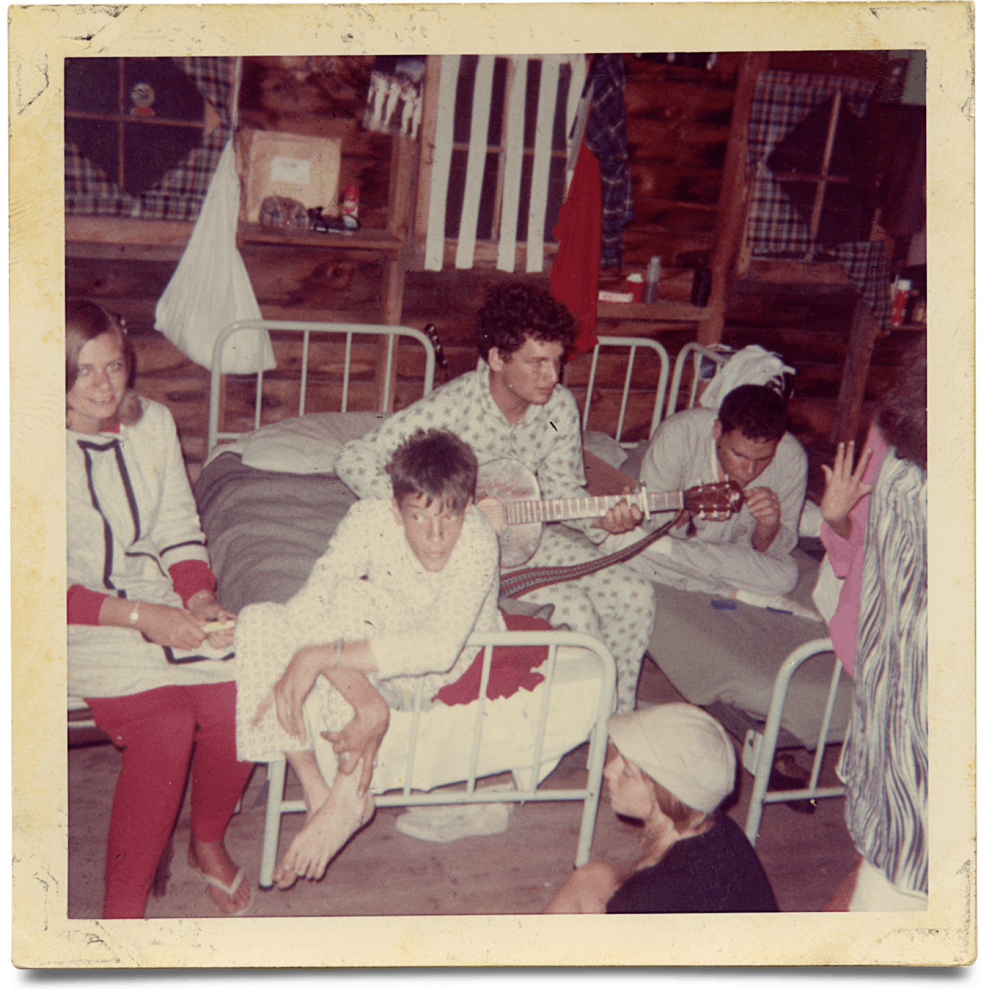 In an old photograph, teens are pictured perched around a cabin in their pajamas. One camper plays guitar. 