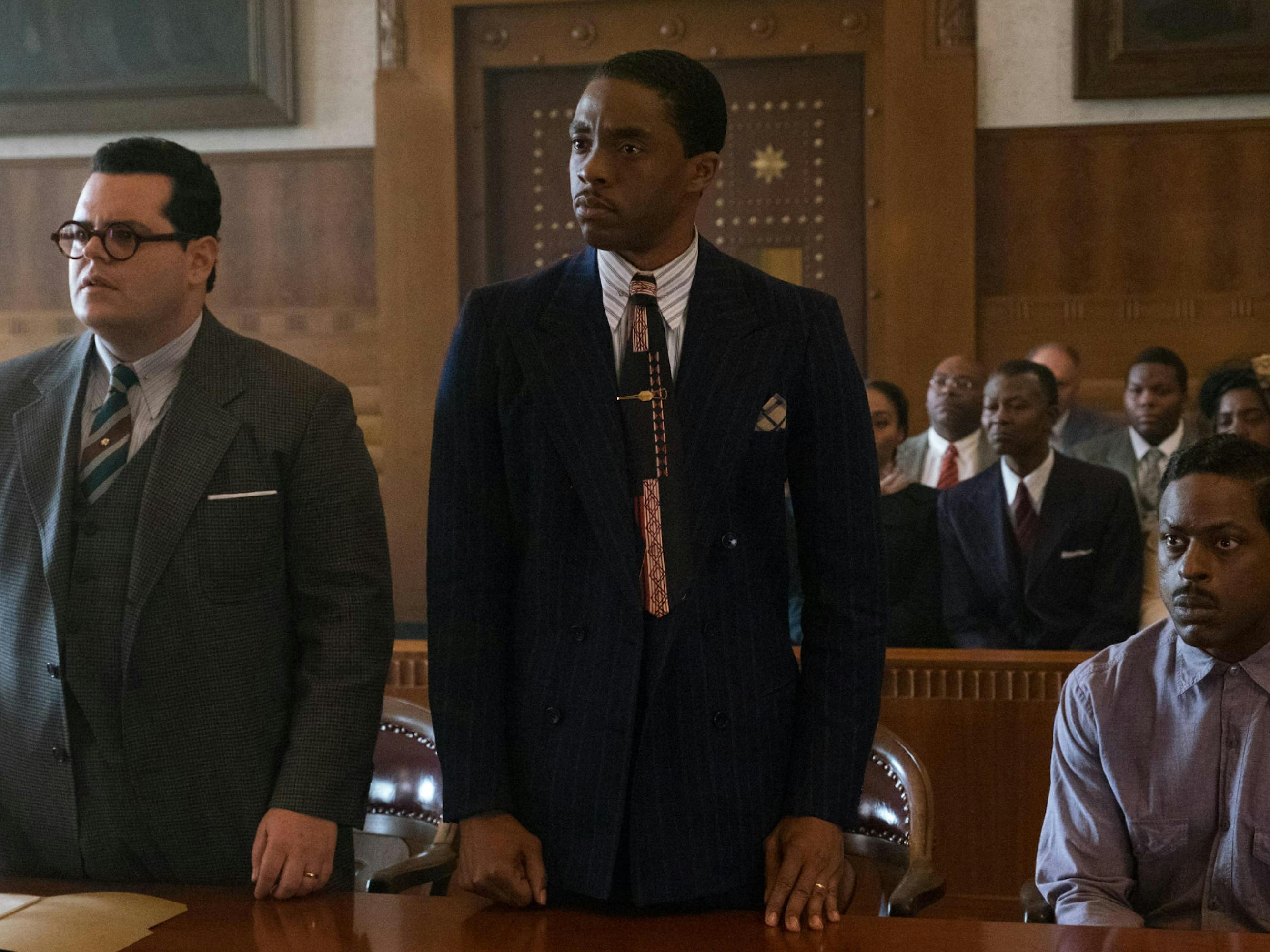 Boseman as Thurgood Marshall. He wears a navy suit and stands between two other men, one in a purple shirt and one in a grey suit. He stands in a courtroom filled with many people and looks off camera with a serious expression.