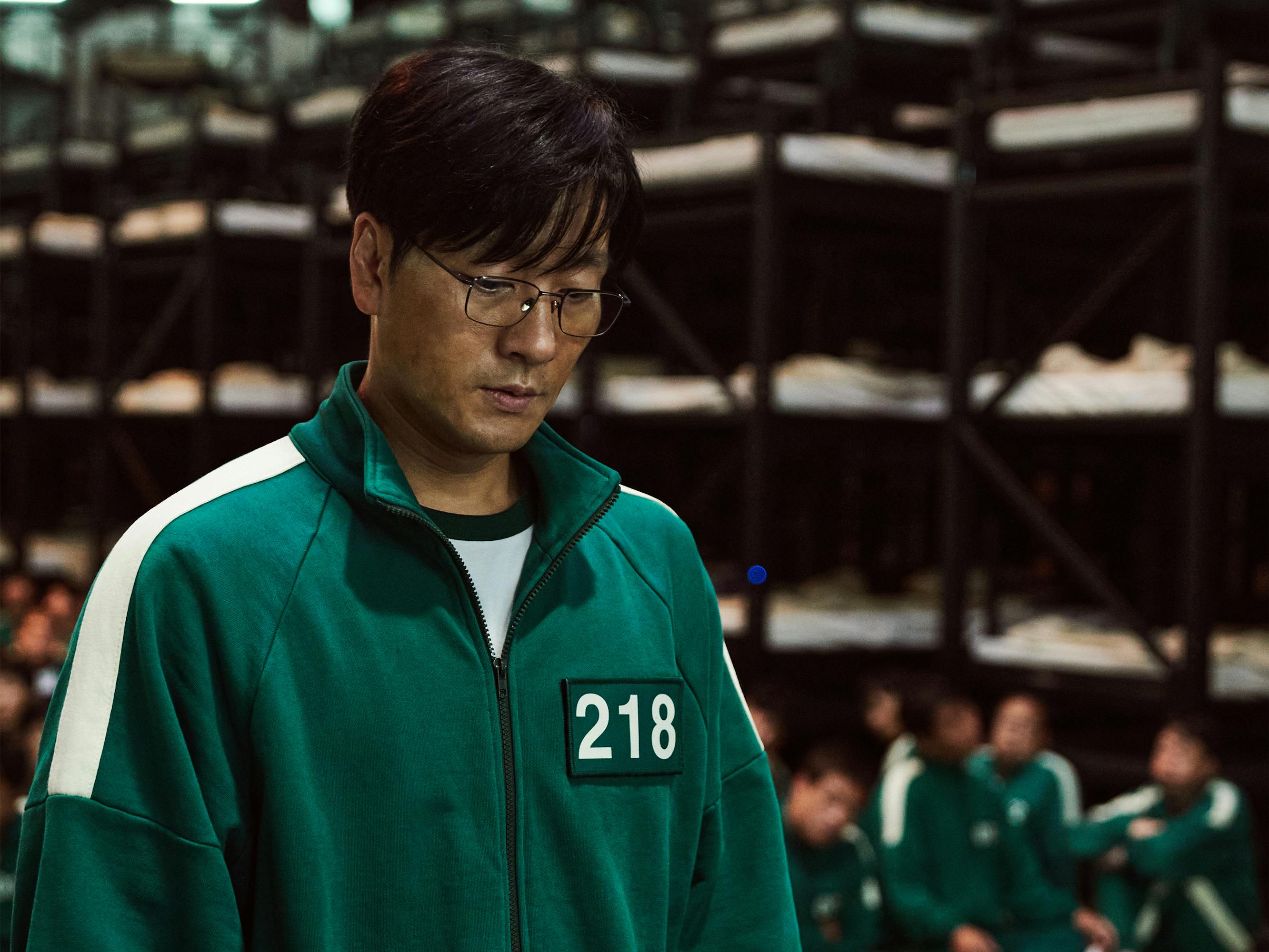 Cho Sang-woo (Park Hae-soo) wears his green tracksuit with 218 and glasses.Behind him are tons of beds and other people in green tracksuits.
