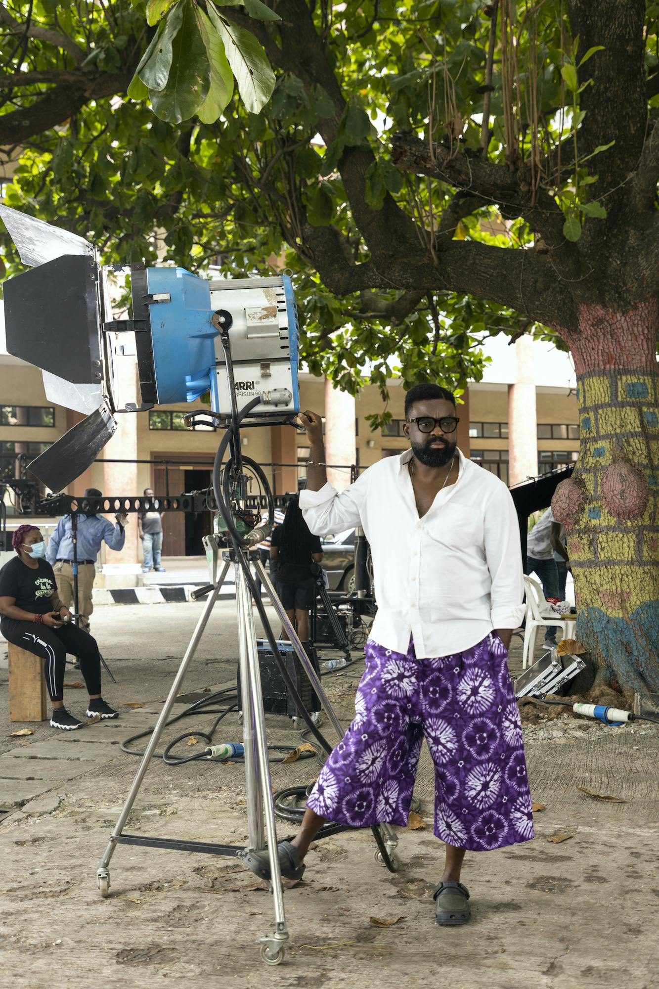 Kunle Afolayan wears a white shirt and purple patterned pants as he films outside. Behind him are masked crew members, equipment, and a leafy tree.