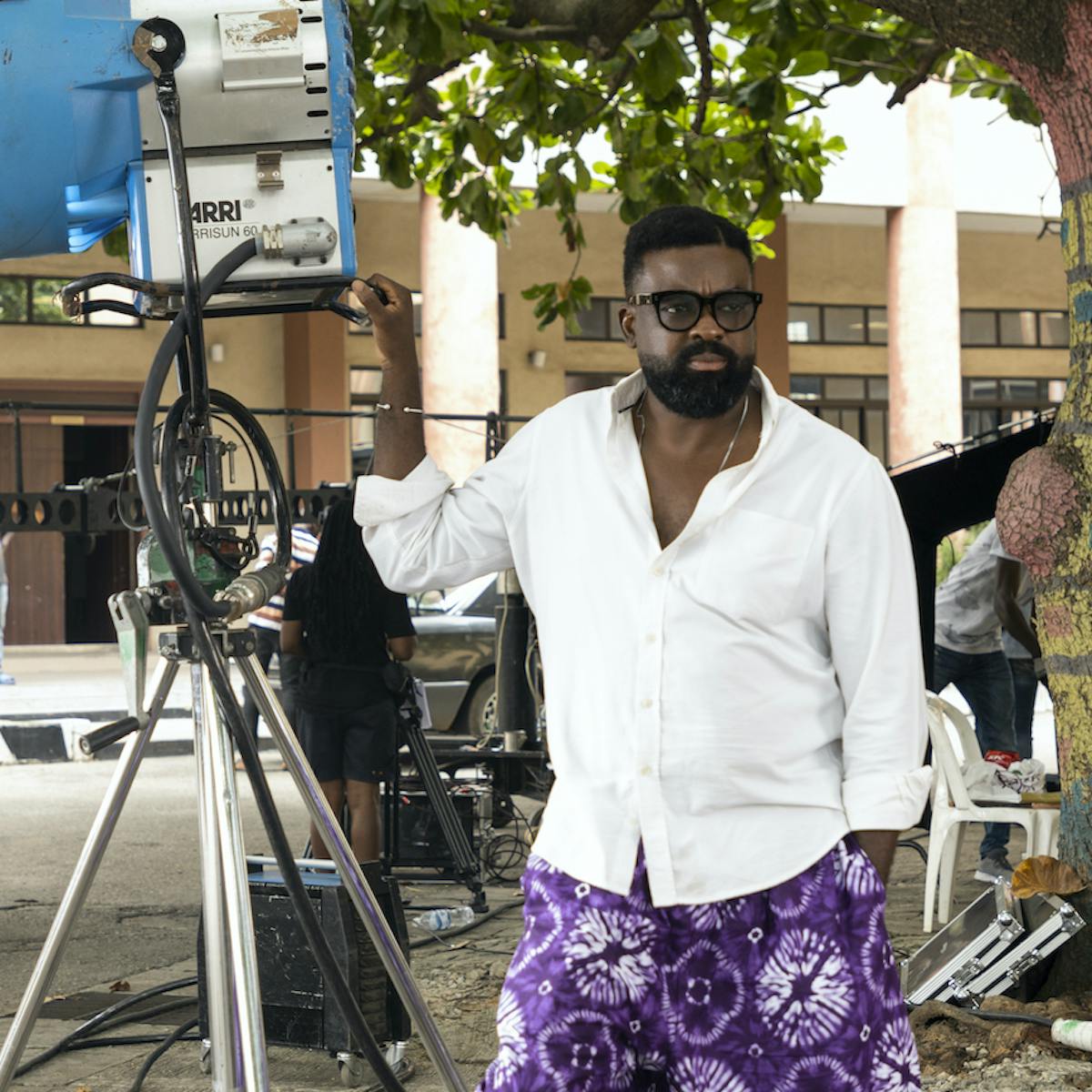 Kunle Afolayan wears a white shirt and purple patterned pants as he films outside. Behind him are masked crew members, equipment, and a leafy tree.