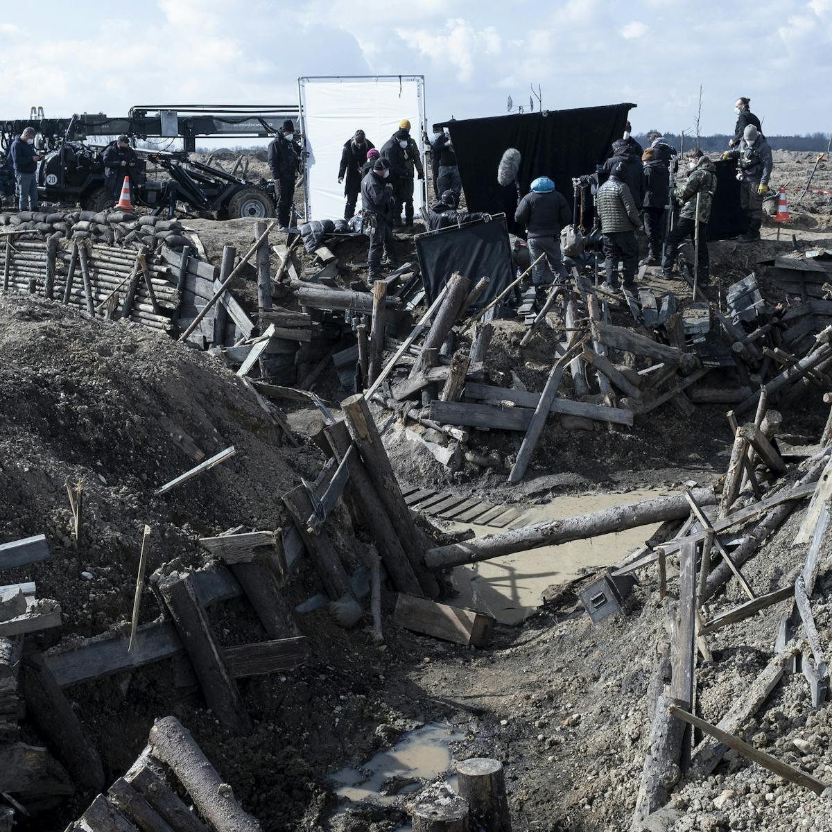 A shot of the trenches, destroyed. Crew members mingle about the film equipment and behind them is a cloudy blue sky. 