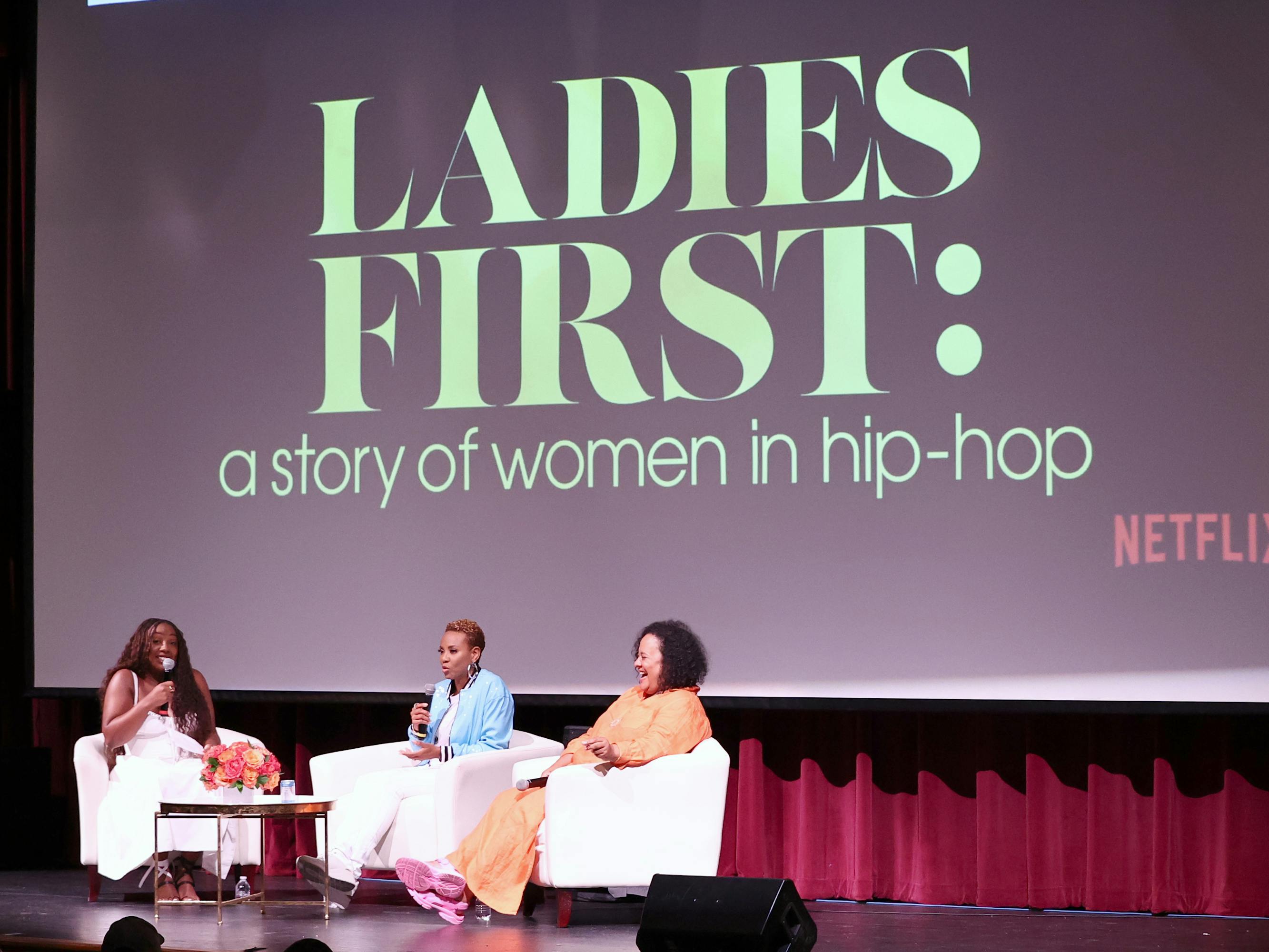 Three women sit on a stage in conversation in front of a projection of the series title "LADIES FIRST: a story of women in hip-hop"