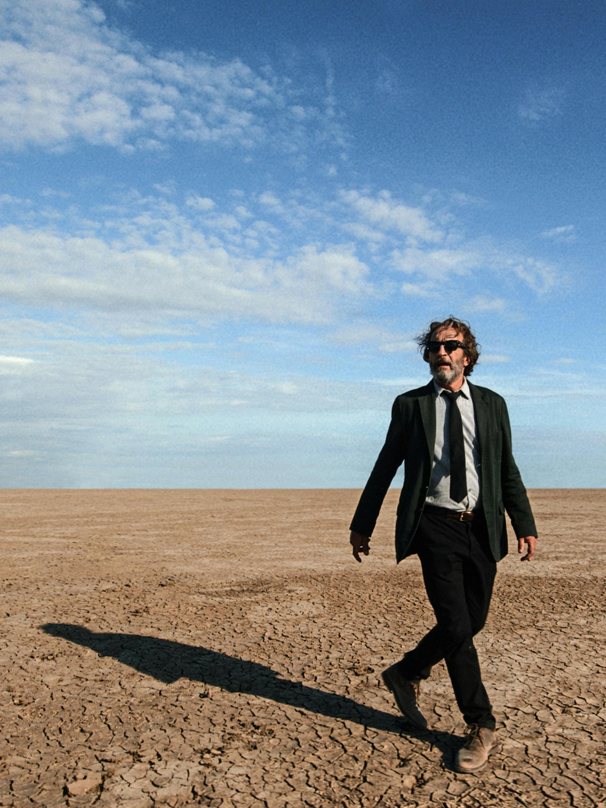 Alejandro González Iñárritu walks on a dusty expanse against a blue sky.