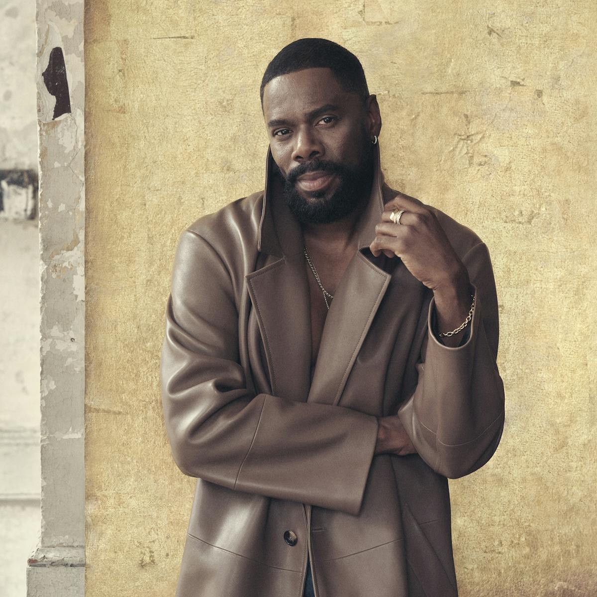 Colman Domingo wears a brown trench coat and stands in front of a gold wall.