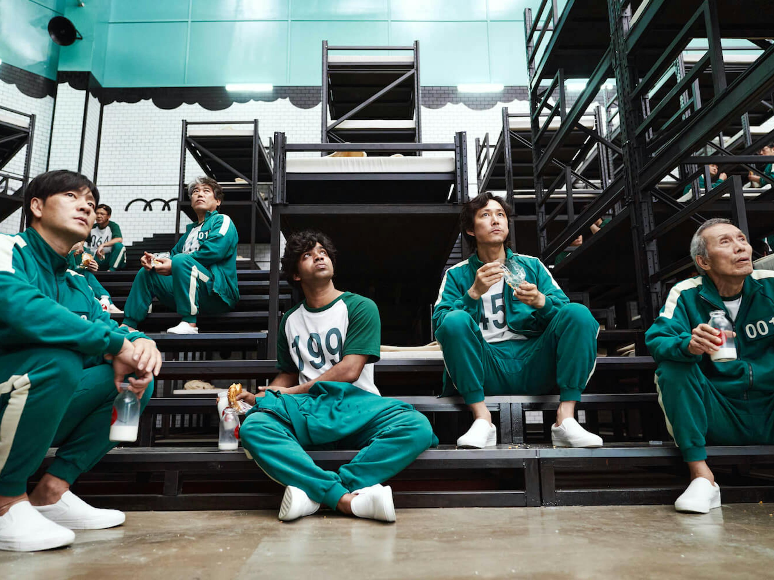 6 contestants sit on the bleachers eating food and drinking milk from jars. The bleachers are dark and behind them are white and teal tiled walls.