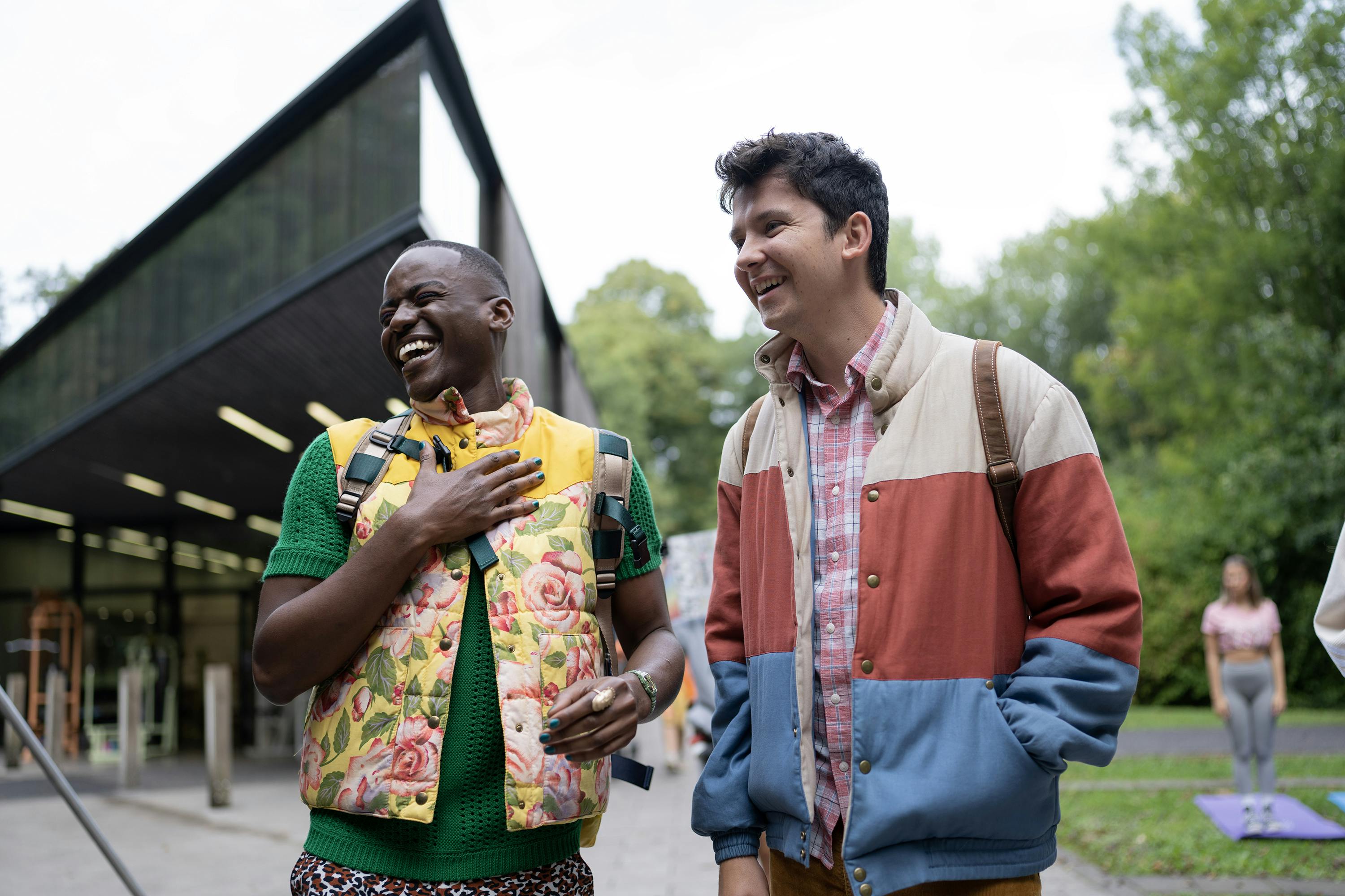 Eric Effiong (Ncuti Gatwa) and Otis Milburn (Asa Butterfield) stand outside their school laughing. Eric wears a flowery puffy vest and Otis wears his trademark white, red, and blue jacket.