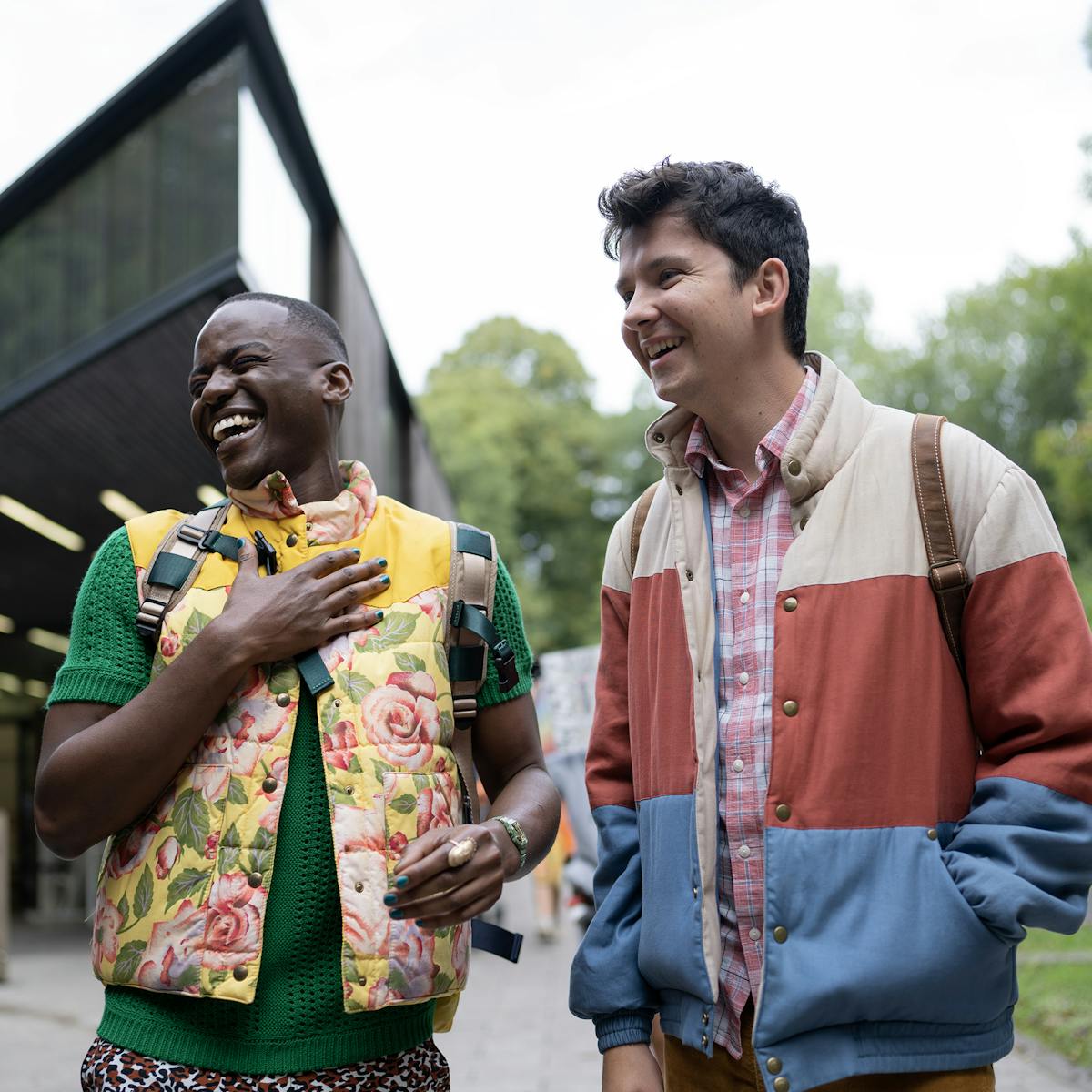 Eric Effiong (Ncuti Gatwa) and Otis Milburn (Asa Butterfield) stand outside their school laughing. Eric wears a flowery puffy vest and Otis wears his trademark white, red, and blue jacket.