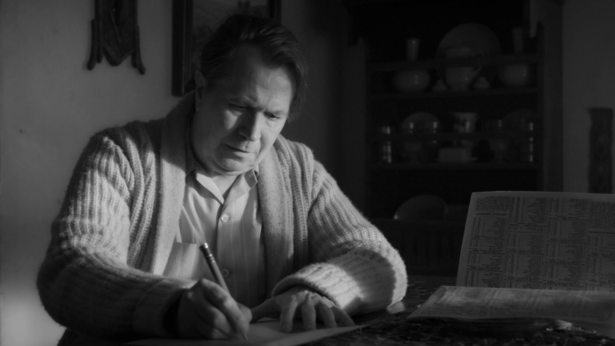 Mank writes at a desk in his Bungalow hideaway in Victorville, California. He wears a cable-knit sweater and his hair falls to one side. Behind him is the rustic decor of the Bungalow: a shelf bearing dishes, etc.; a small framed painting hanging on one of the unevenly textured white walls.