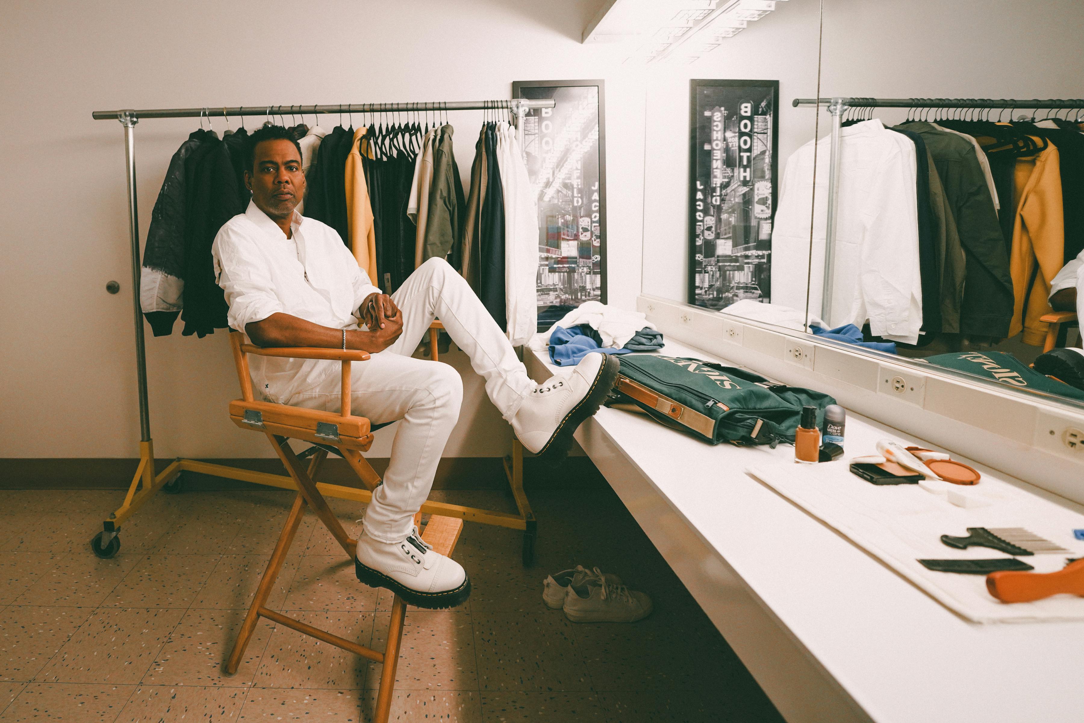 Chris Rock wears a white shirt, khaki pants, and white shoes in his dressing room. 
