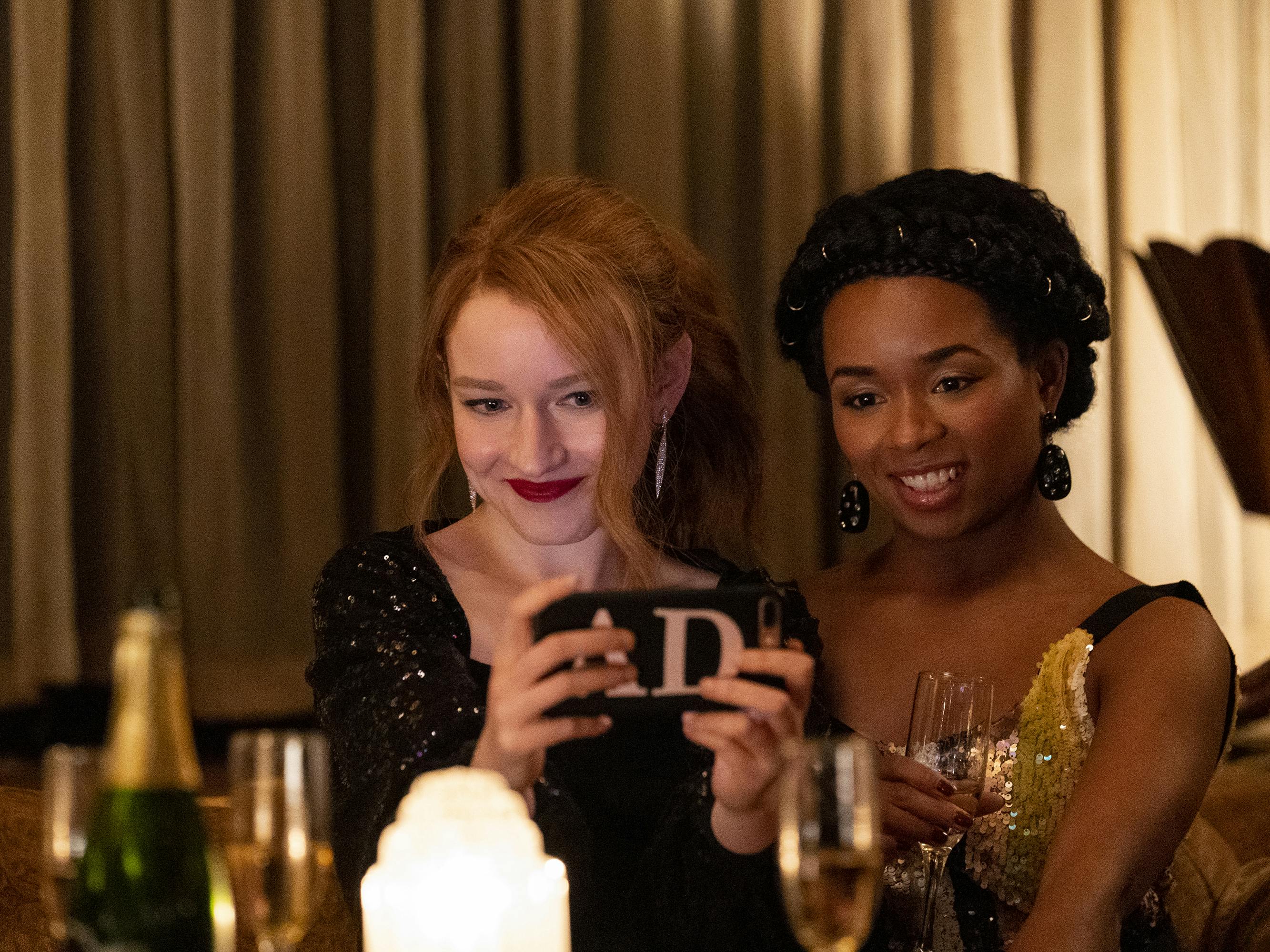Julia Garner as Anna Delvy and Alexis Floyd as Neff Davis sit in a bar and pose for a selfie while surrounded by champagne bottles and flutes.