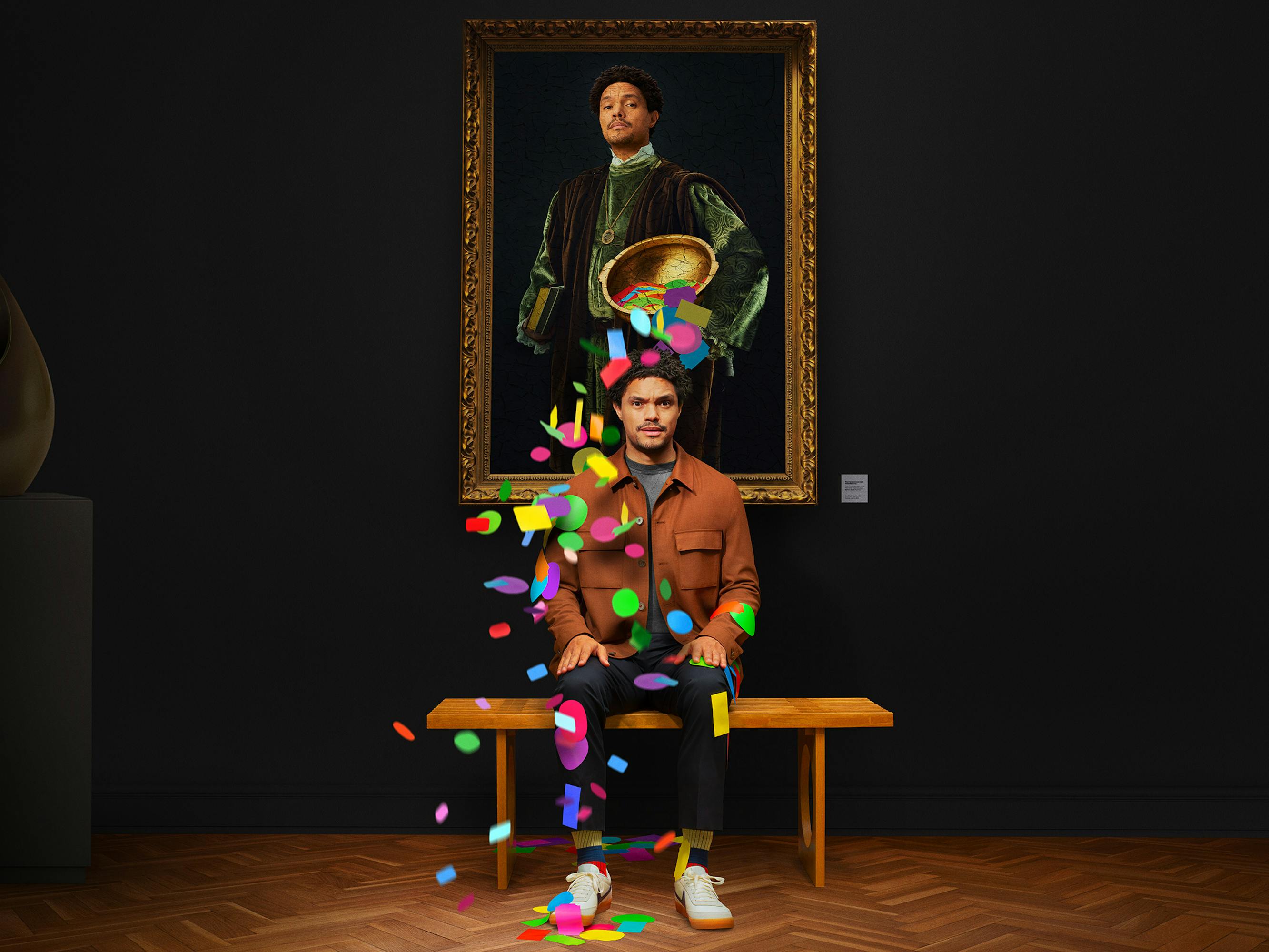 Trevor Noah wears a brown jacket and sits in front of a portrait of himself, as a bucket of stickers falls over his head.