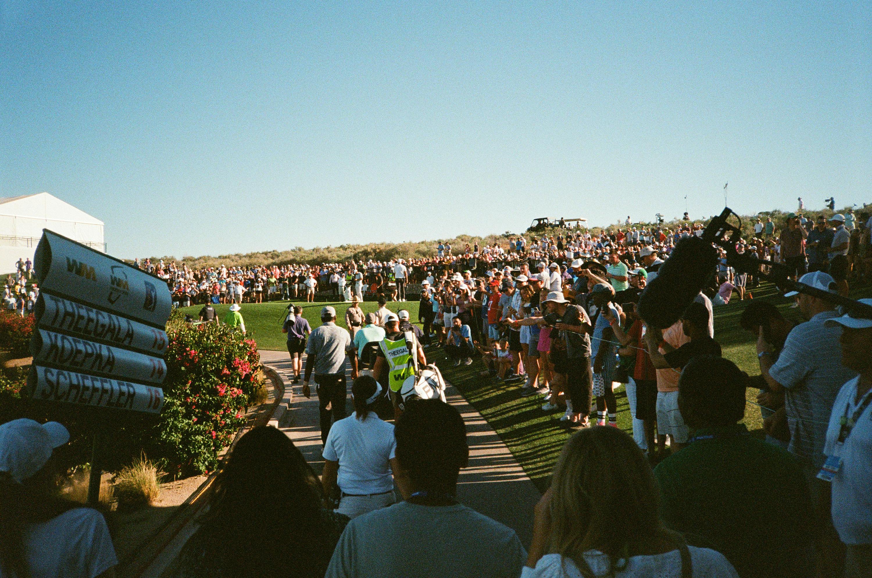 Some golfers walk through a crowd of spectators on a sunny day. 