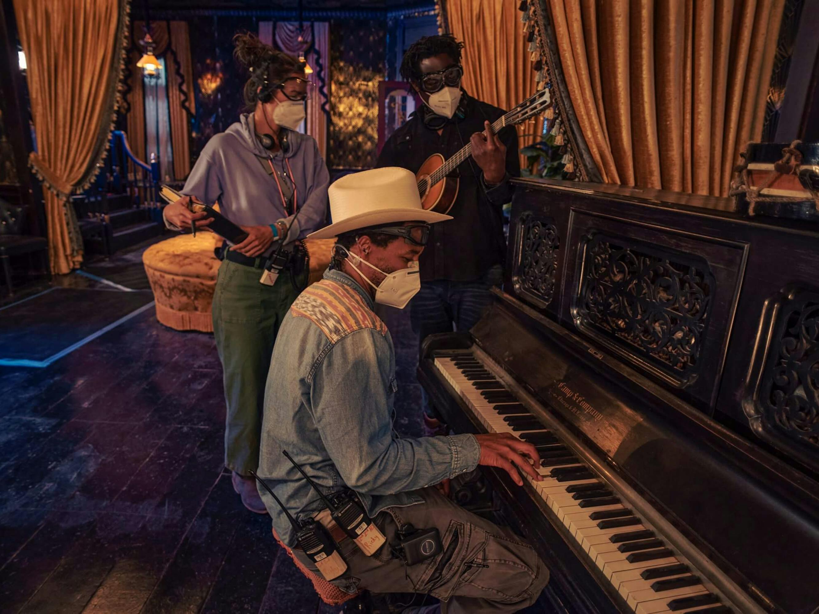 Jeymes Samuel and The Harder They Fall crew stand around a piano. Samuel plays a bango, one of the crew members plays a piano, and the lsat carries a billboard.