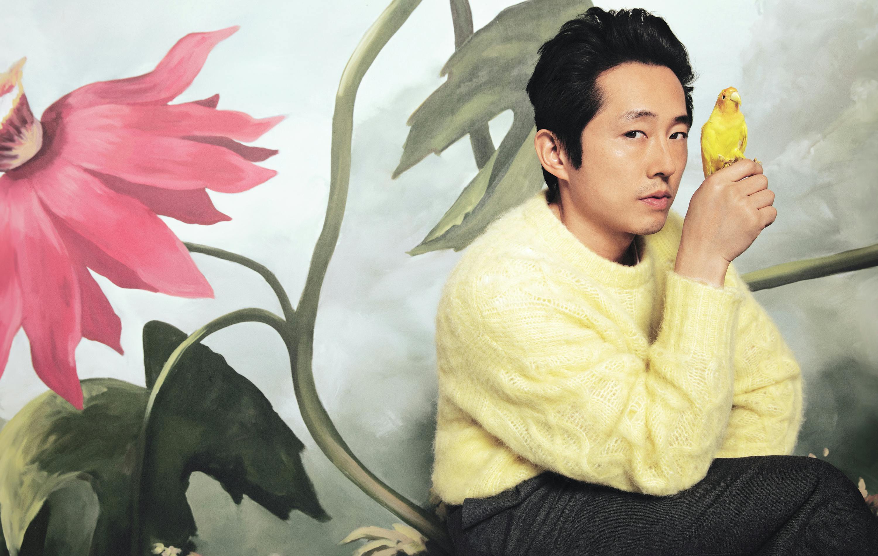 Steven Yeun wears a yellow sweater and black pants. A little yellow bird is perched on his hand. Behind him are some painted flowers on the wall. 