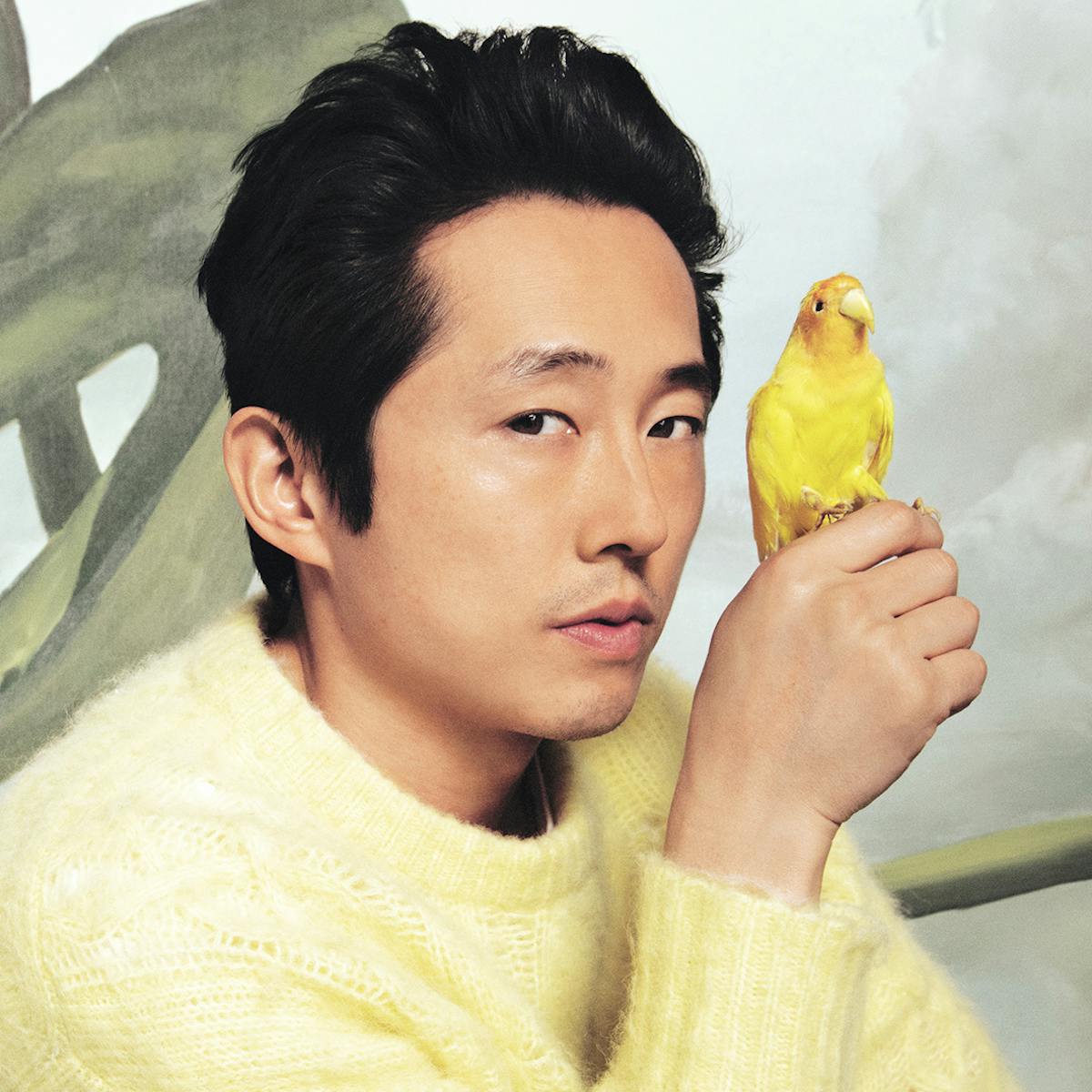 Steven Yeun wears a yellow sweater and black pants. A little yellow bird is perched on his hand. Behind him are some painted flowers on the wall. 