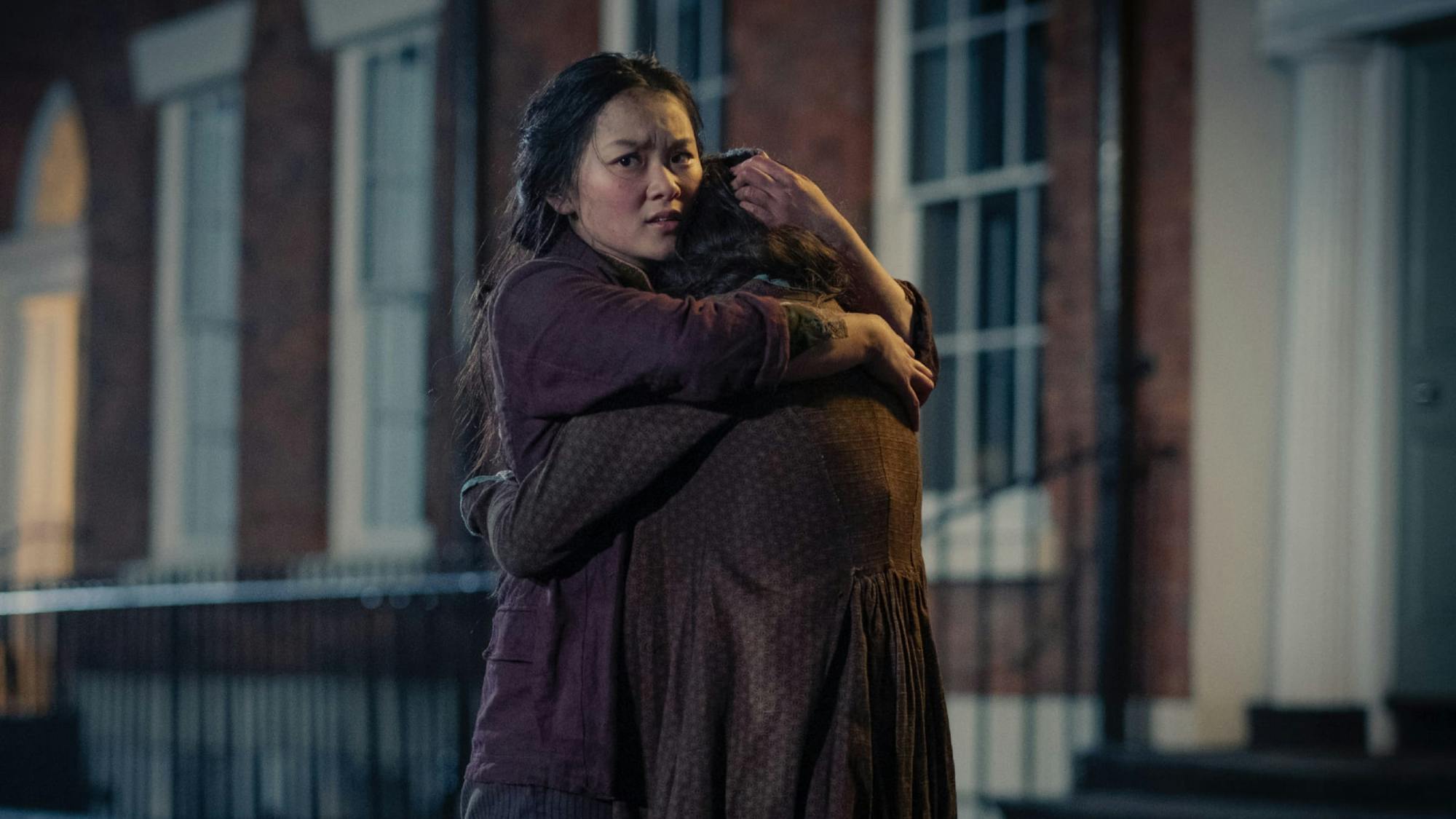 Standing in front of a row of townhouses in evening light, Bea embraces Jessie. She stares out over Jessie’s shoulder, looking worried.