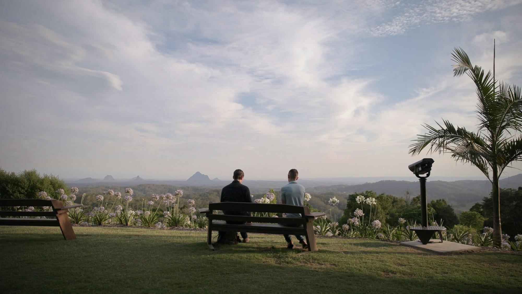 Two people sit on a bench in an idyllic spot with flowers, palm trees, and a blue sky.