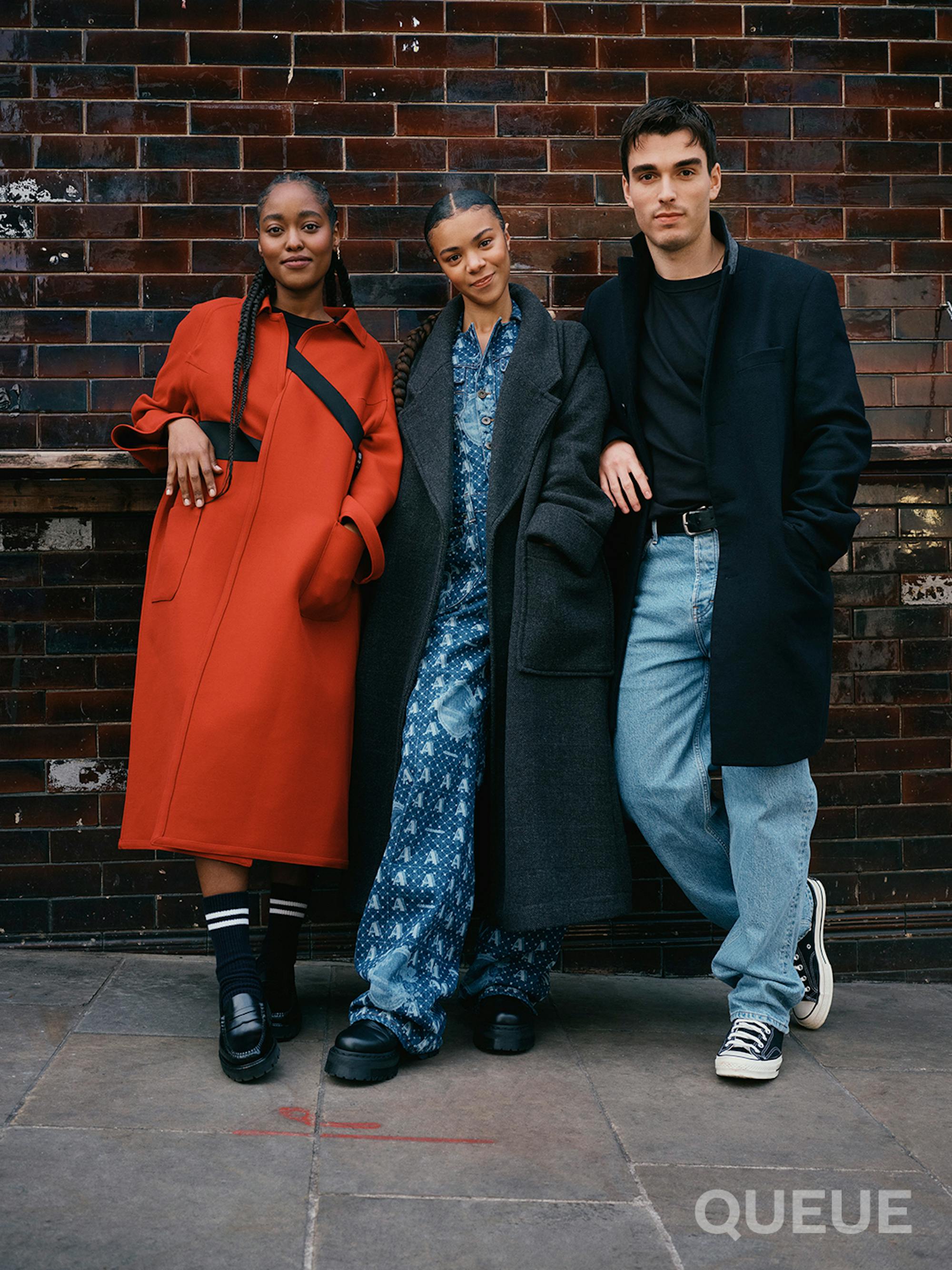 Arsema Thomas, India Amarteifio, and Corey Mylchreest stand together against a brick wall. Thomas wears a red jacket. Amarteifio wears a grey jacket atop a blue jumpsuit. Mylchreest wears a black jacket over a black tee and lightwash jeans.