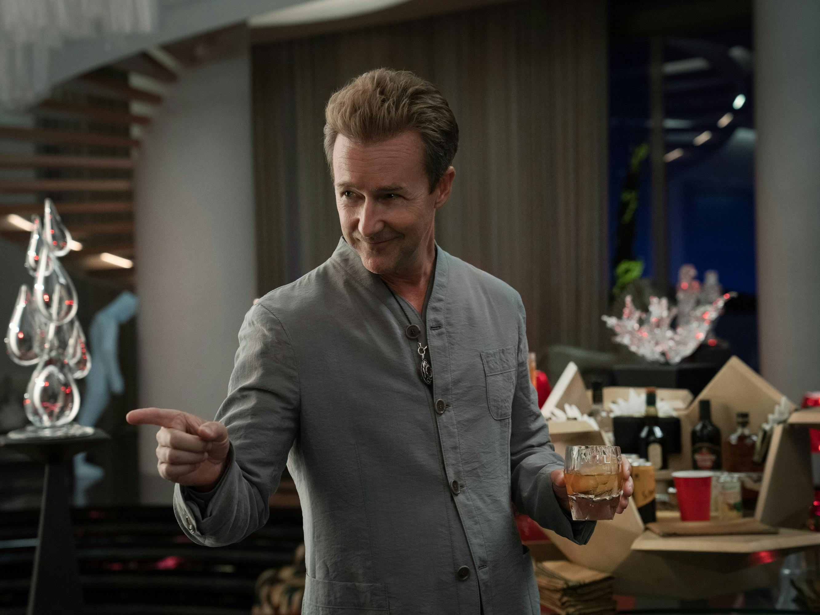 Miles Bron (Edward Norton) wears a gray top and holds a glass of brown liquor. He points slyly in someones direction.