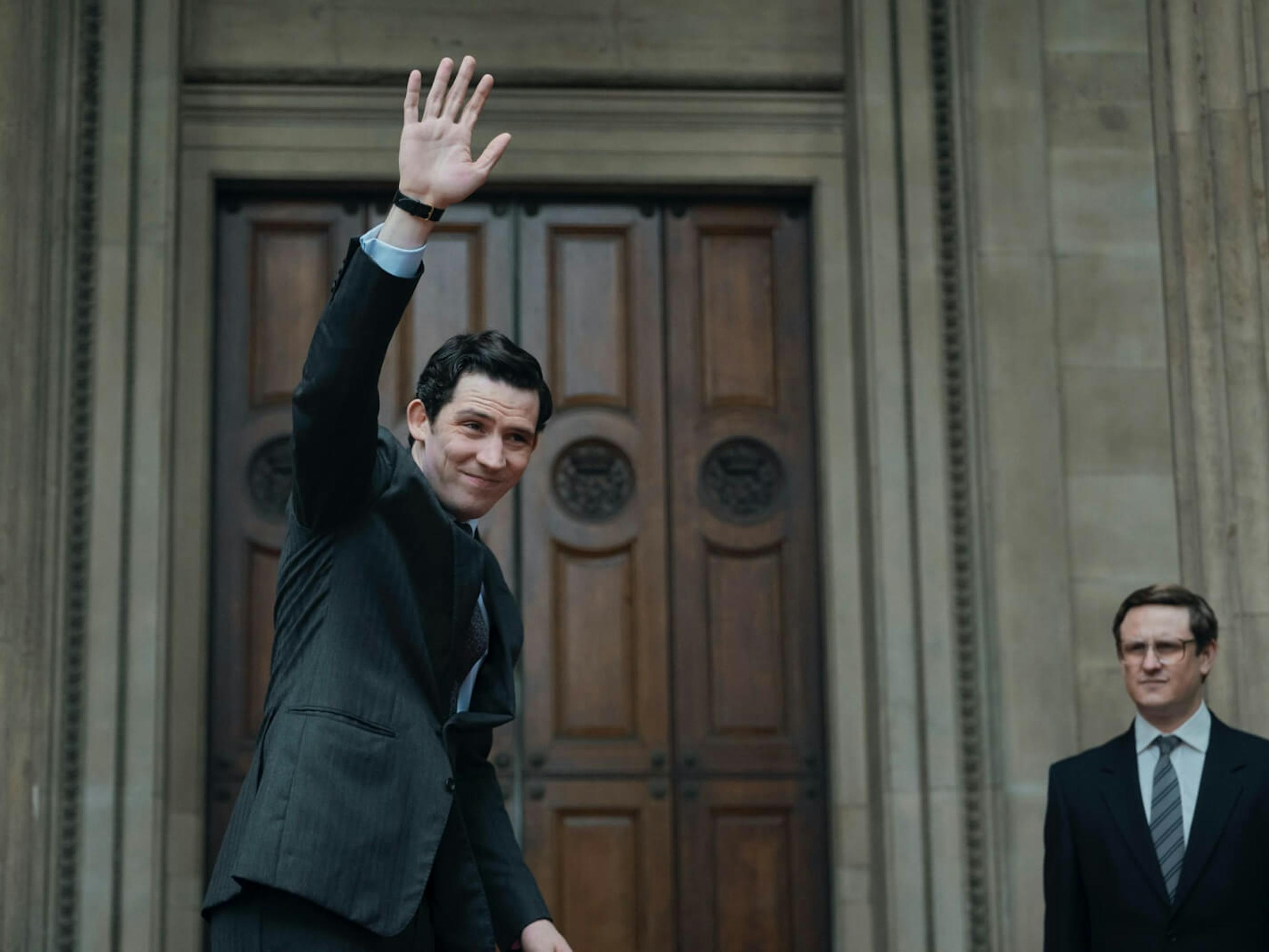Prince Charles (Josh O’Connor) waves in a dark suit. Behind him is another suited man, and an impressive wooden door.