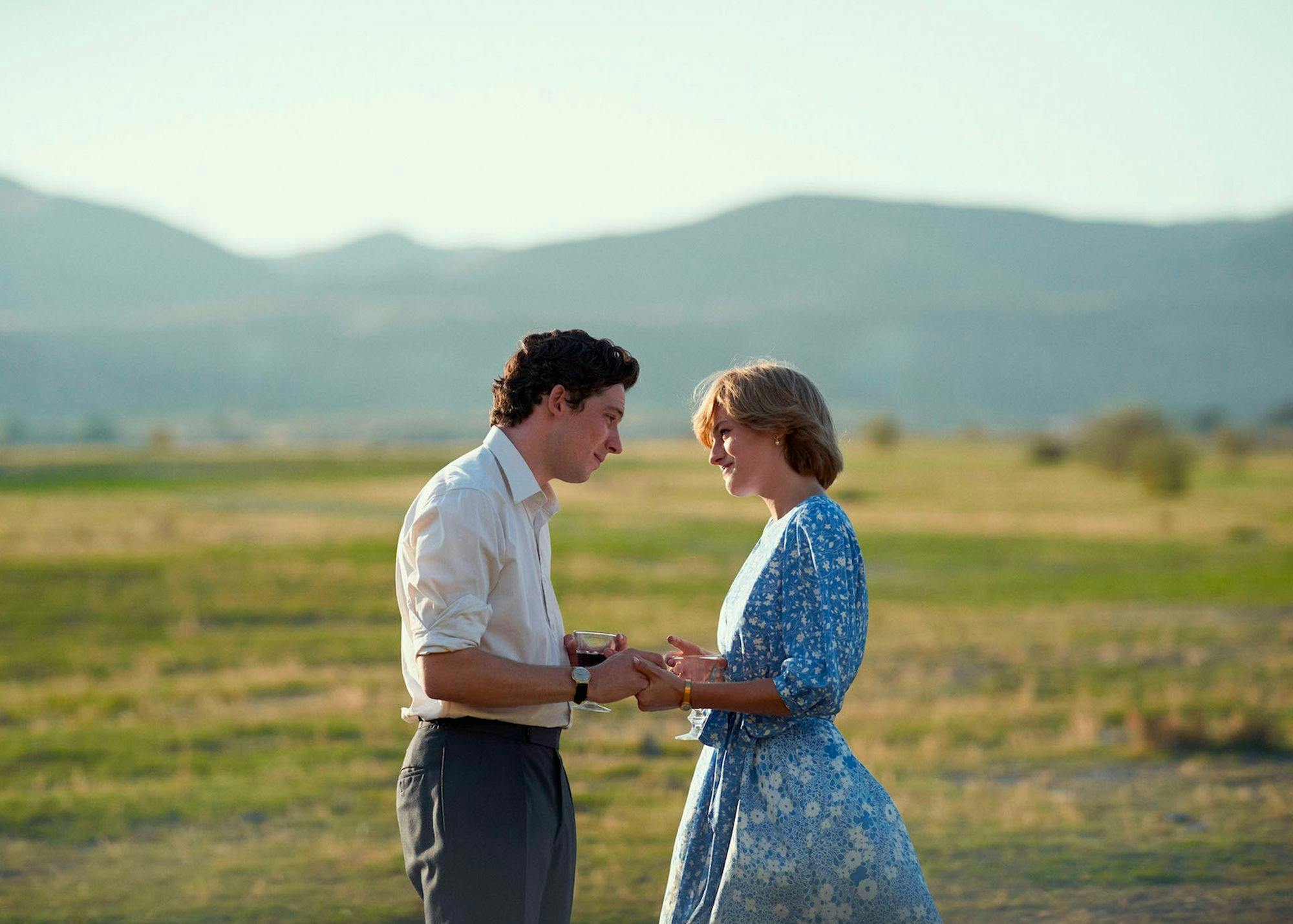 Prince Charles (Josh O’Connor) and Princess Diana (Emma Corrin) hold hands in a sun-lit desert. O’Connor wears dark pants and a white shirt, and Corrin wears a blue flowered dress.