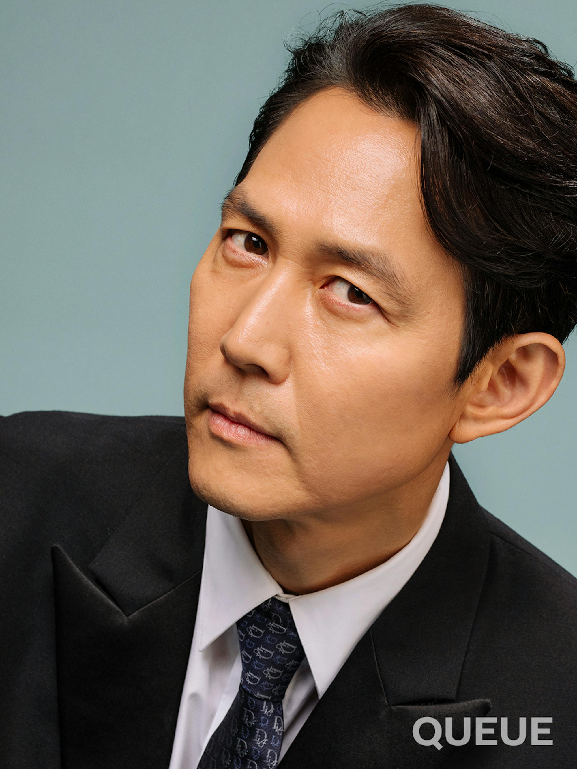 Lee Jung-jae in front of a gray-blue background looking stoic and sophisticated in a suit and tie.