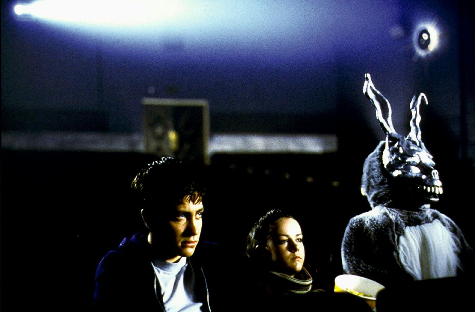 Donnie Darko (Jake Gyllenhaal) and Gretchen Ross (Jena Malone) in Donnie Darko (2001) sit beside the man in a large rabbit suit in a movie theater. The shot is dark but Gyllenhaal looks angry and wearied.