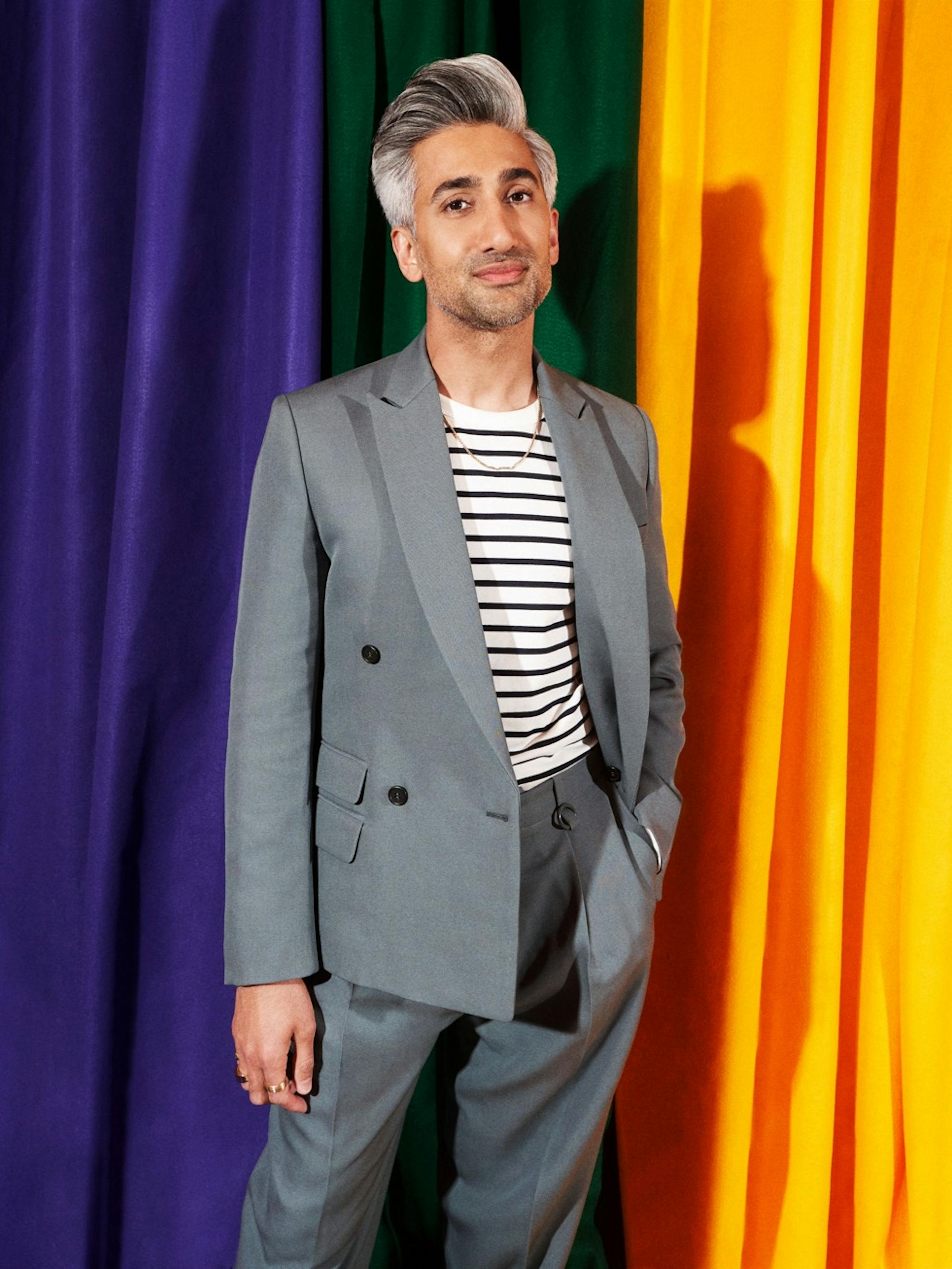 Tan France wears a grey suit, white and black striped shirt. His hair is coiffed. Behind him are purple, green, and yellow hanging cloths.