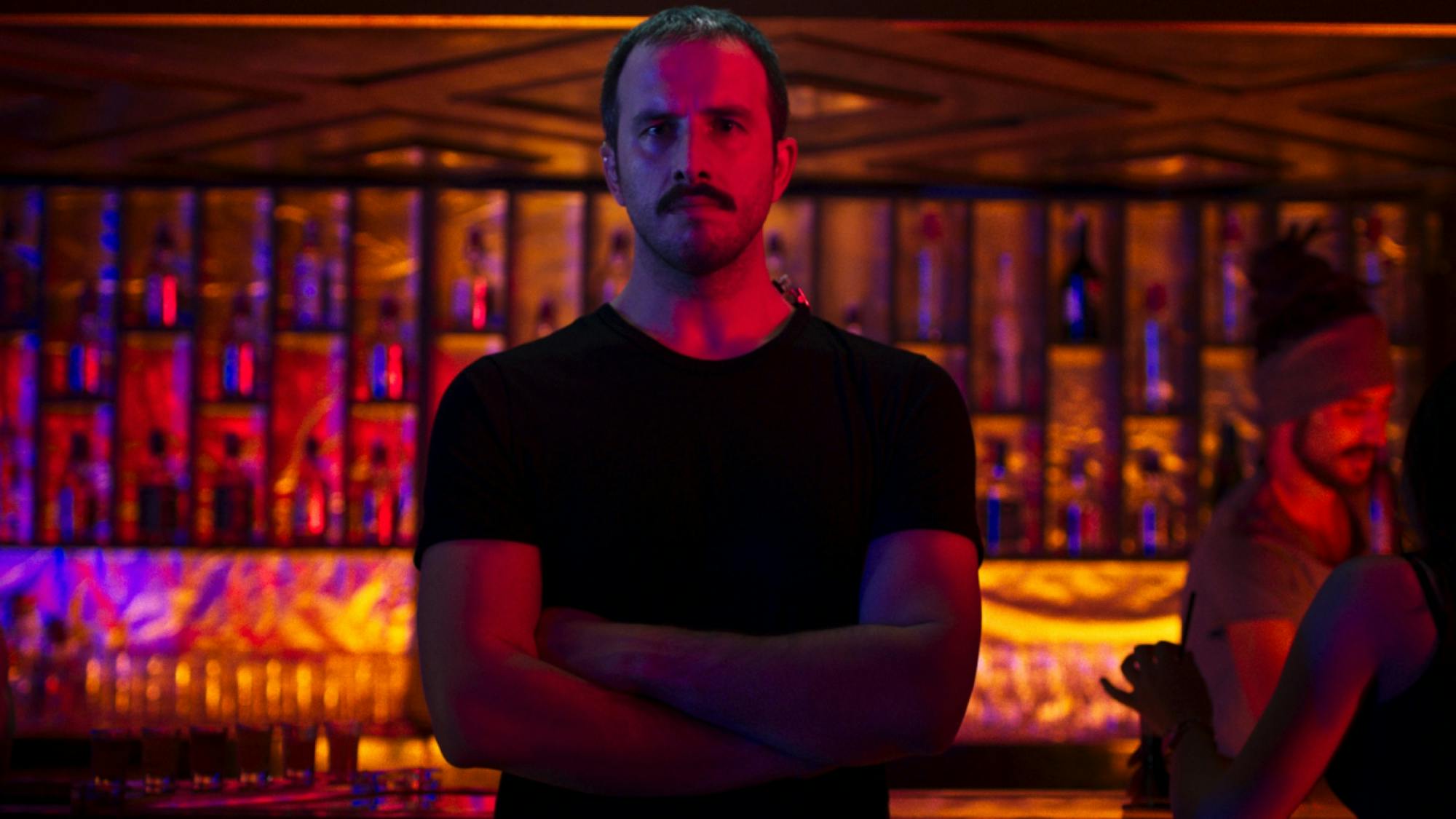 Yasin stands in front of a fashionable bar, arms crossed over a black t-shirt. The scene is cast in red, orange, and purple light.