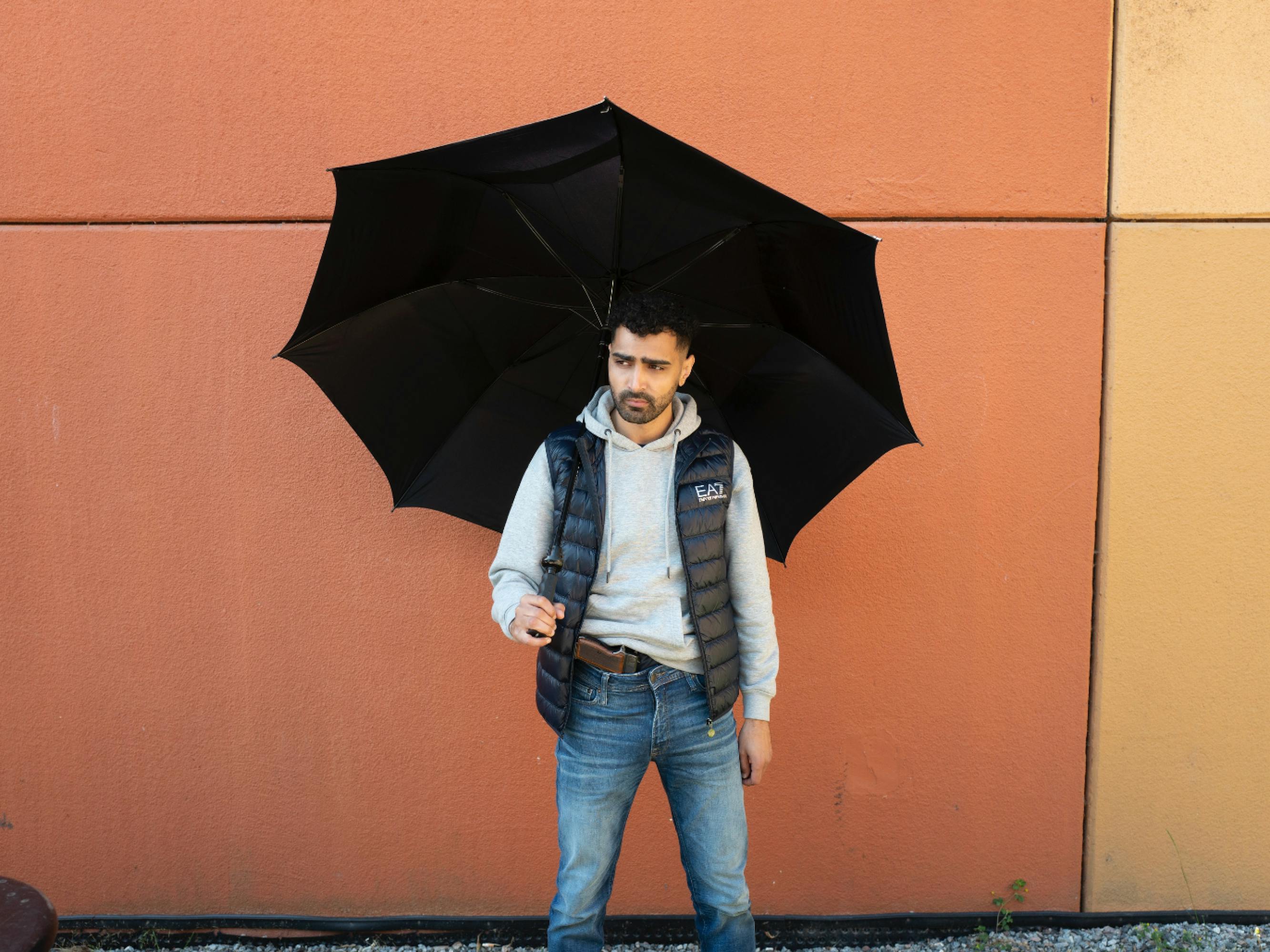 Alexander Abdallah wears jeans, a grey hoodie, and a black puffer vest. There is a gun in his front pocket, and he holds an umbrella above his head. There is an orange wall behind him.