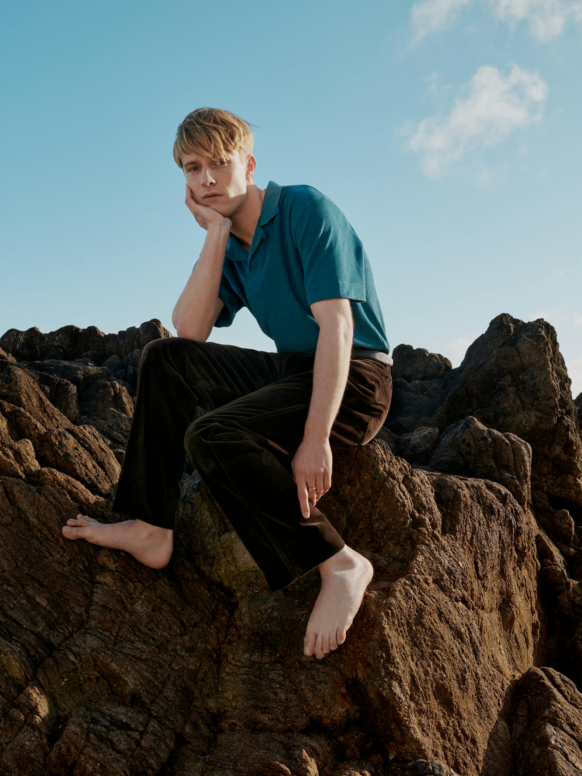 Louis Hofmann wears brown pants and a teal polo and sits barefoot on a rock.
