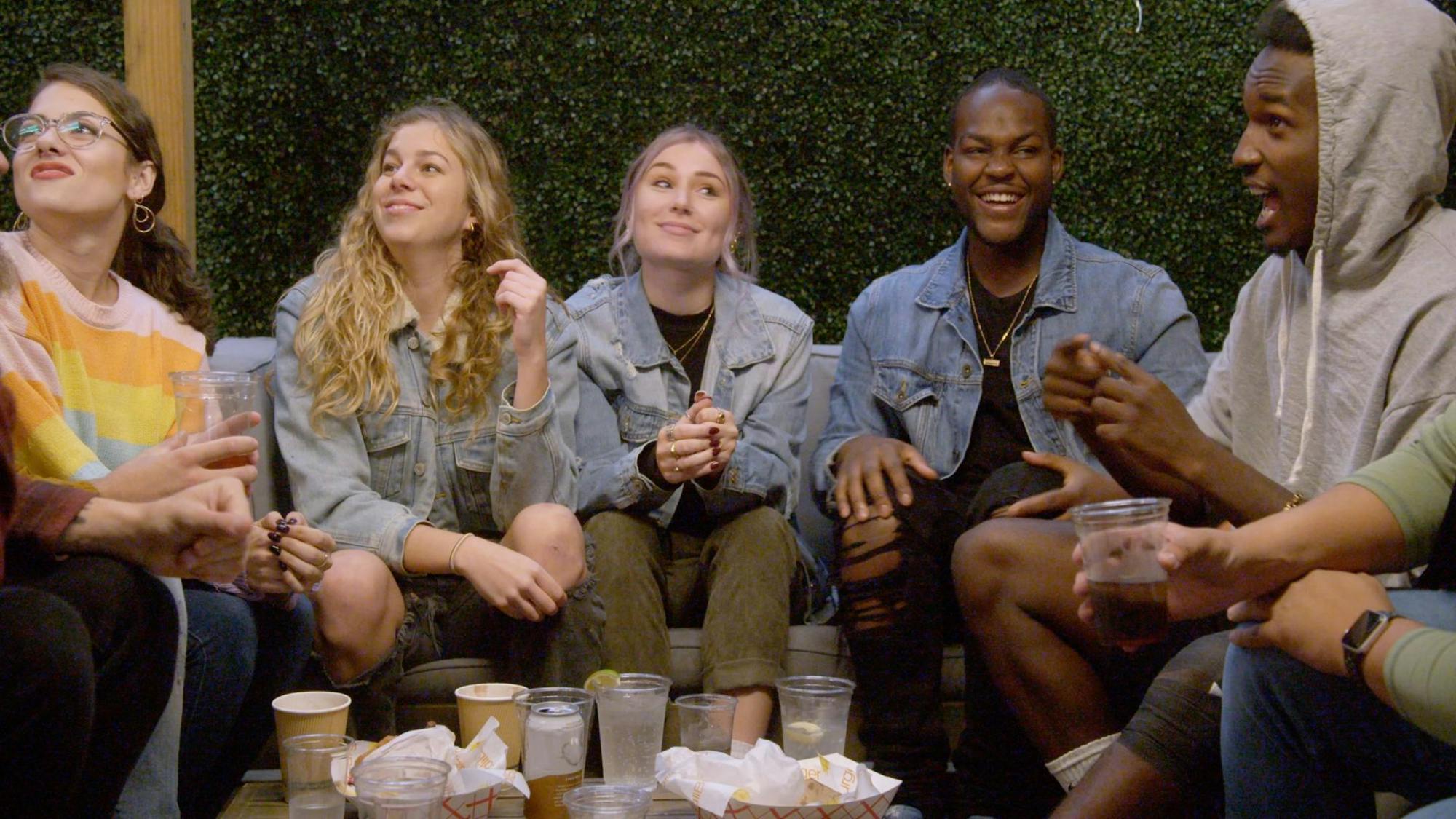 5 students sit on a couch around a table. From left to right: Renate Rose, Alexa Paulay-Simmons, Cheyenna Clearbrook, Daequan Taylor, and Rodney Burford There are coffee and water cups scattered in front of them, as well as containers of fries. Rose wears a striped sweater, Paulay-Simmons, Clearbrook, and Taylor wear jean jackets, and Burford wears a greya hoodie. Taylor is laughing at something Burford is saying, and Paulay-Simmons and Rose are looking off camera. The scene is lighthearted and fun!