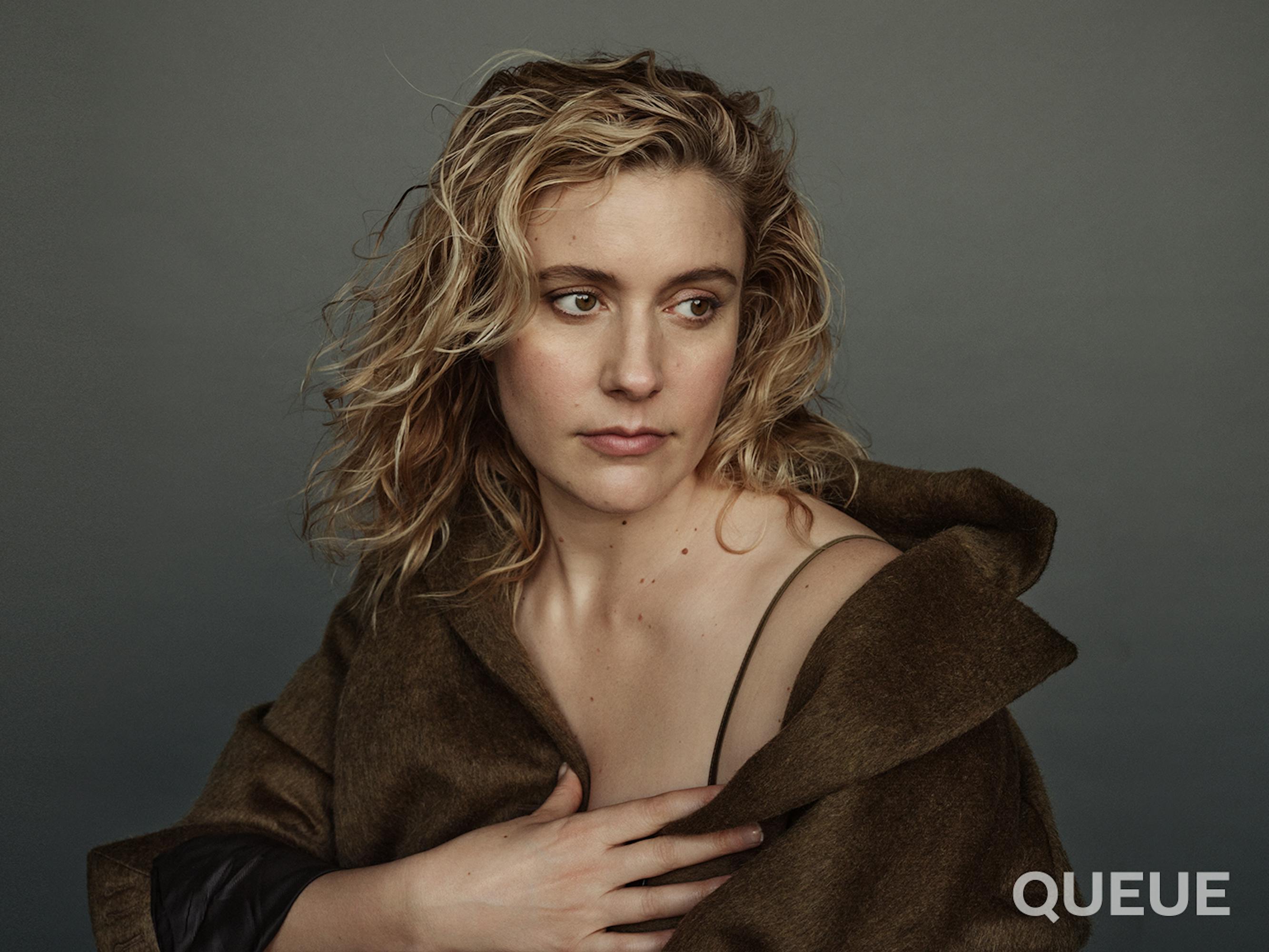 Greta Gerwig wears a brown jacket that reveals a thin a black strap.