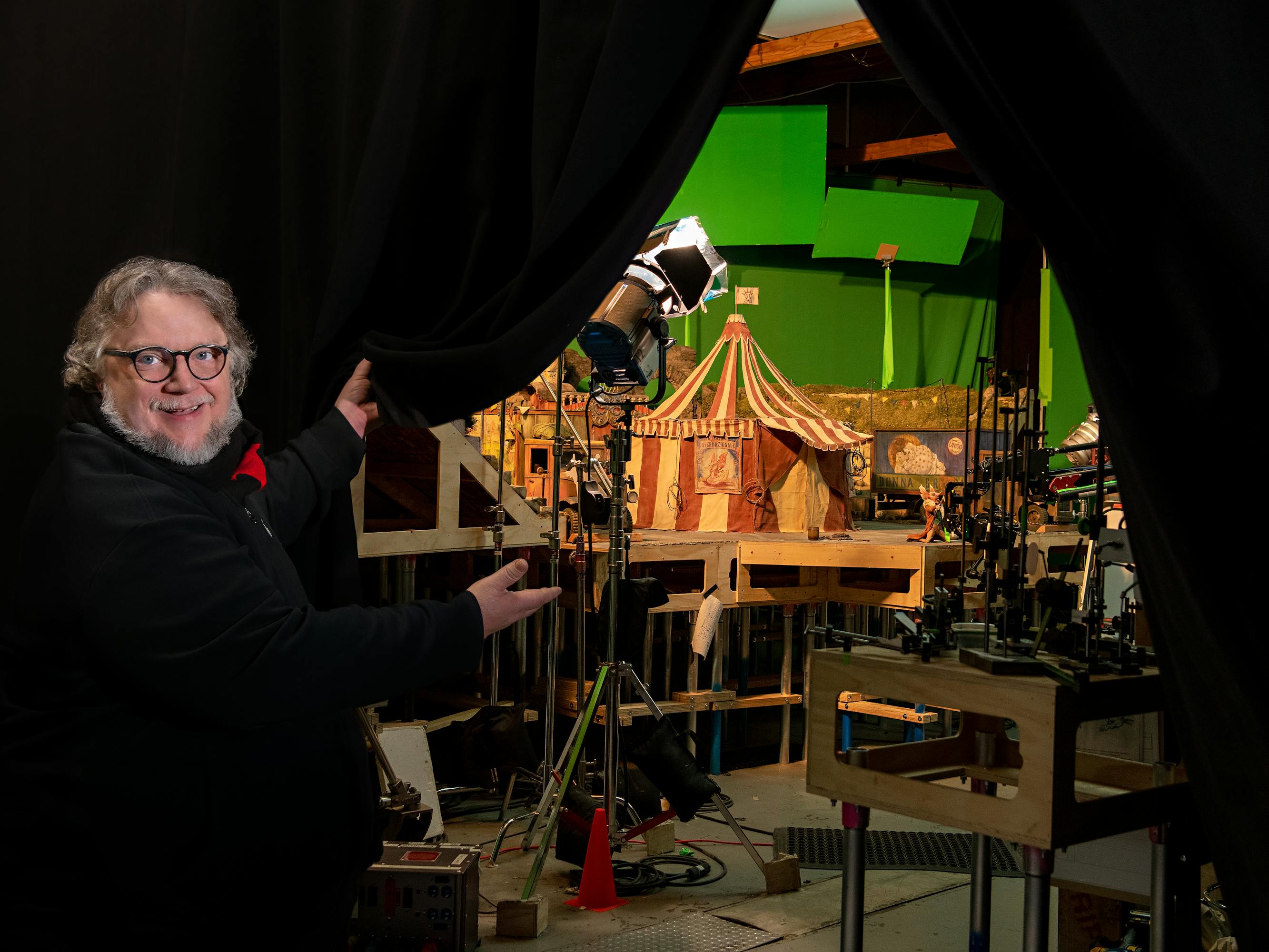 Guillermo del Toro wears all black and glasses and gestures to his set on a small stage set against a green screen.
