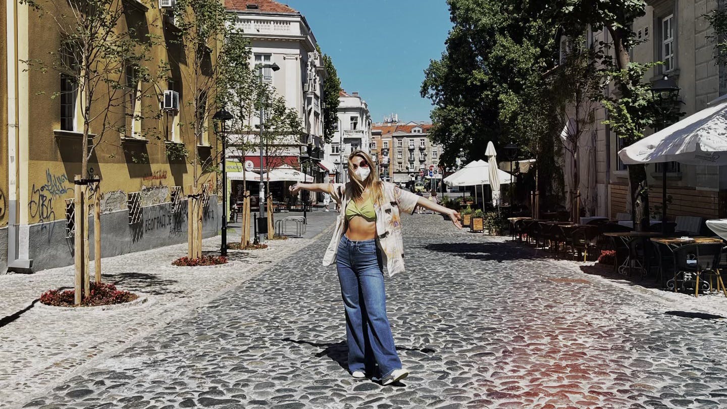 Madelyn Cline wears baggy blue jeans and a green crop top with a mask and sunglasses. She stands in a cobble street on a sunny day.