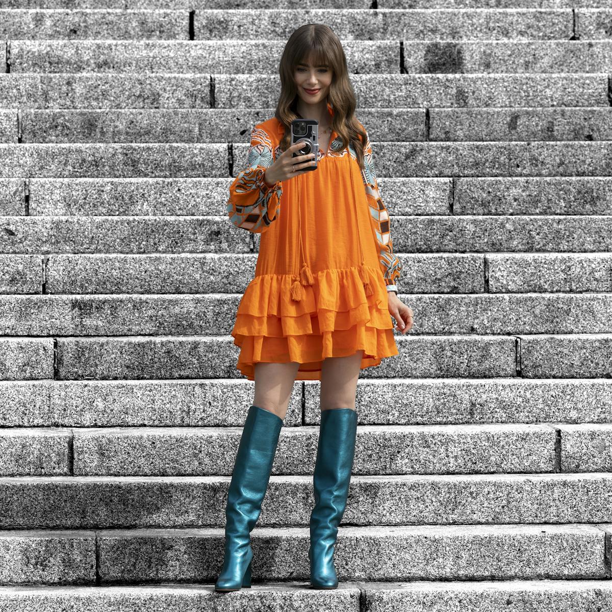 Emily Cooper (Lily Collins) wears an orange dress and blue heeled boots as she takes a selfie on some stone steps.