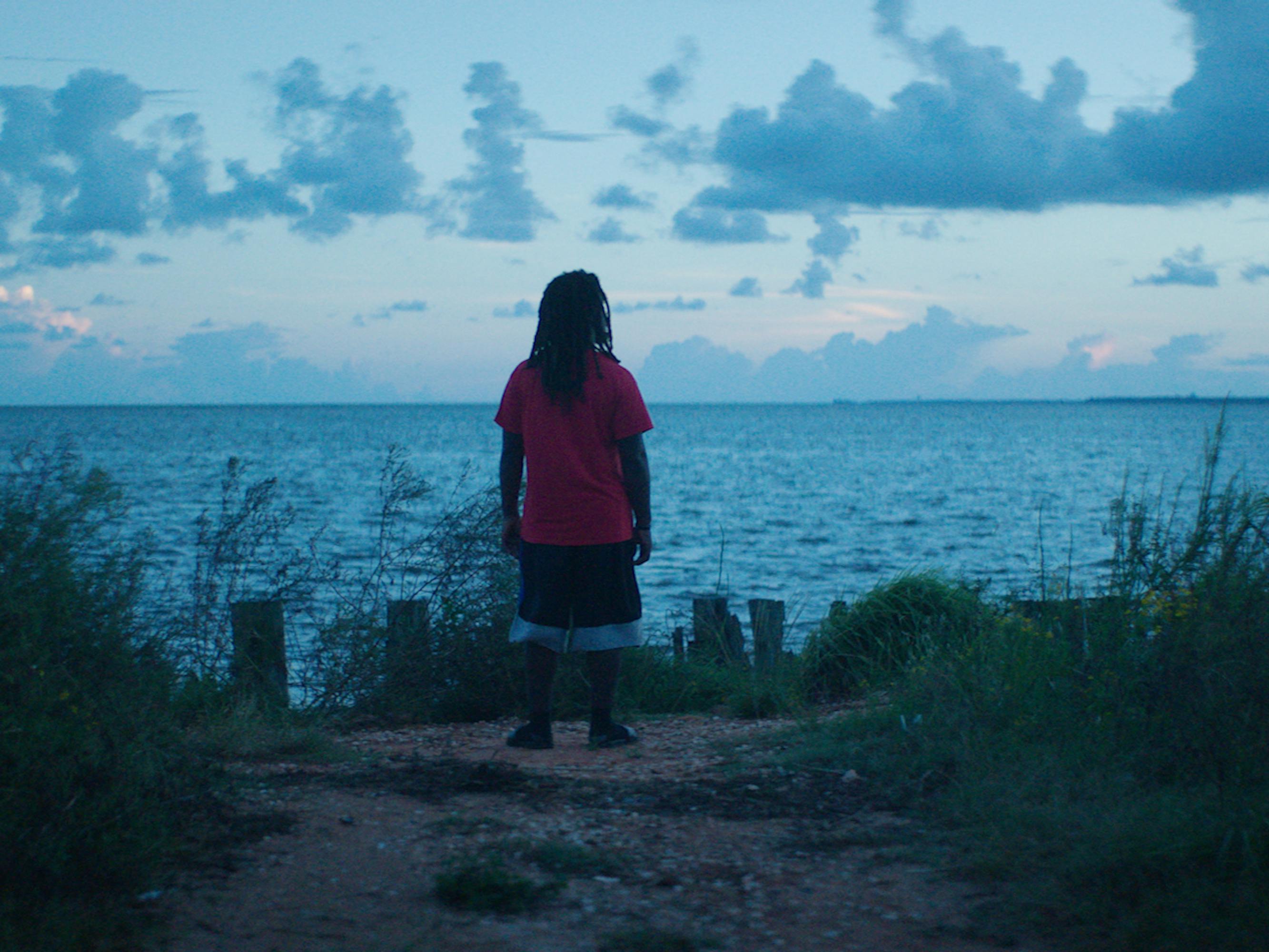 One of the people from Margaret Brown's Descendant stands on a beach at dawn looking out on the turquoise water. 