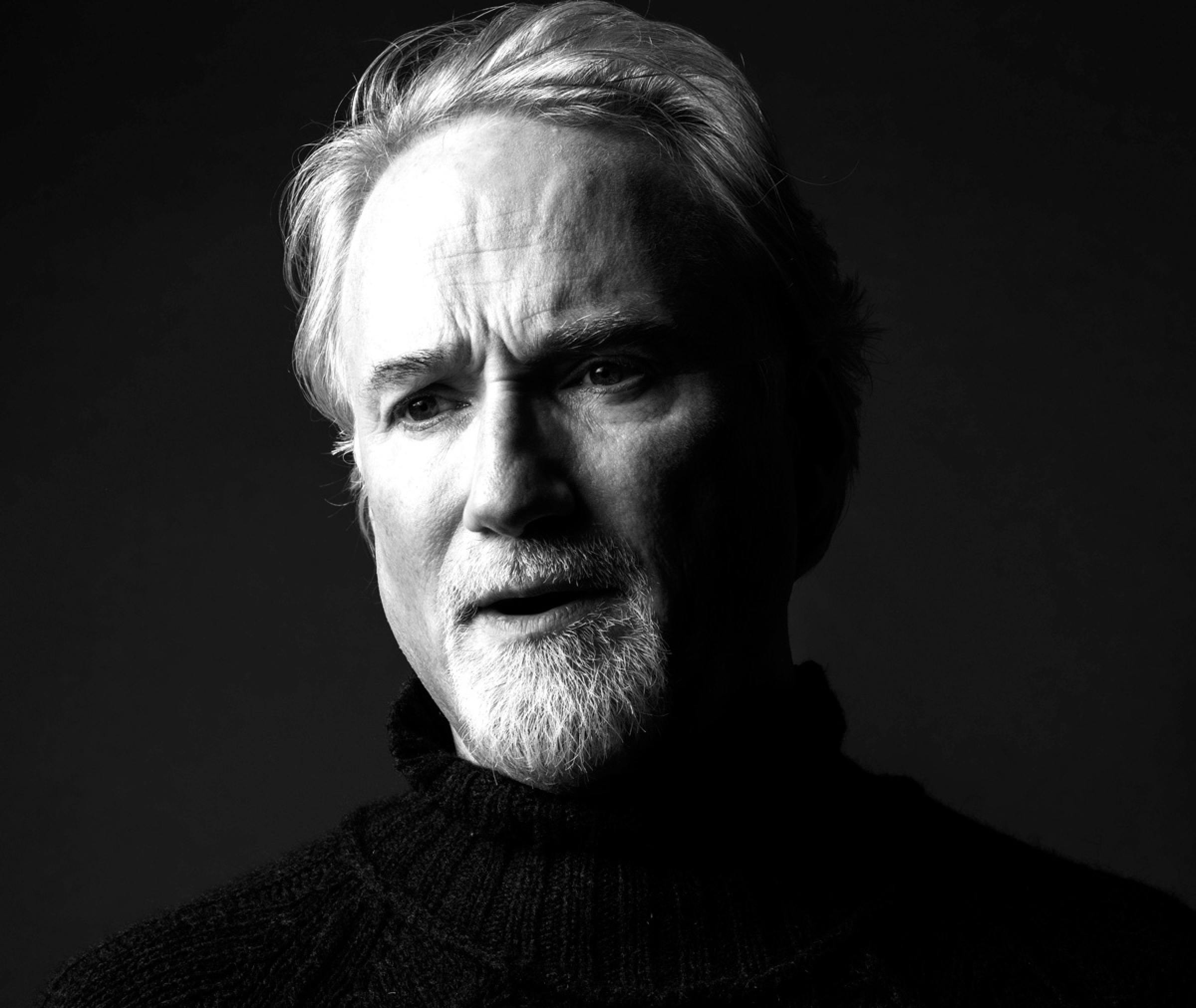 A high-contrast black-and-white portrait of David Fincher. He wears a black turtleneck and looks past the camera.