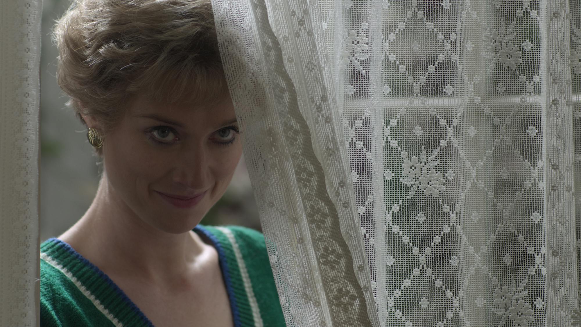 Princess Diana (Elizabeth Debicki) wears a green sweater and peeks out from behind a lacy white curtain.