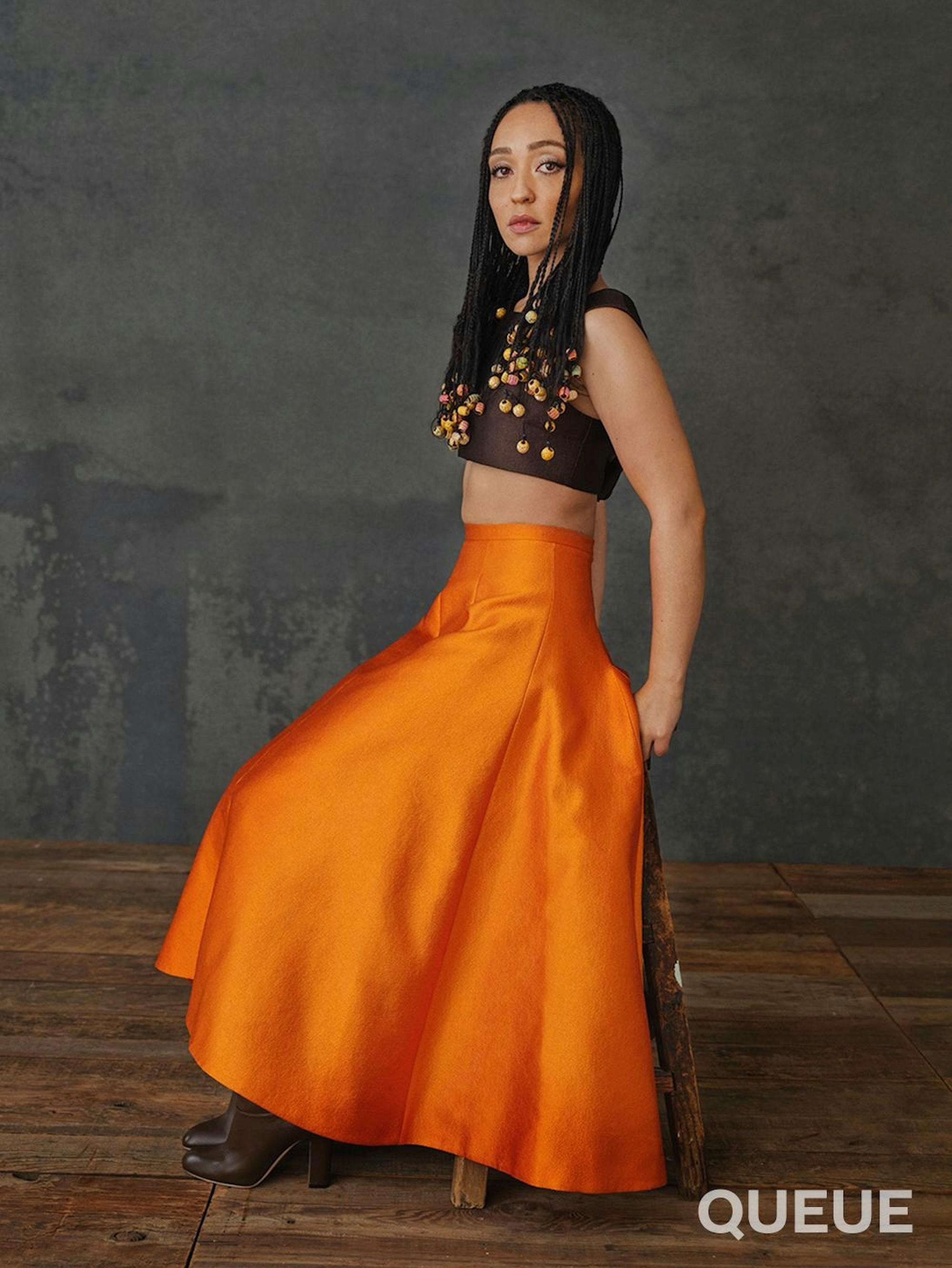 Ruth Negga wears a long orange skirt and brown crop top. 