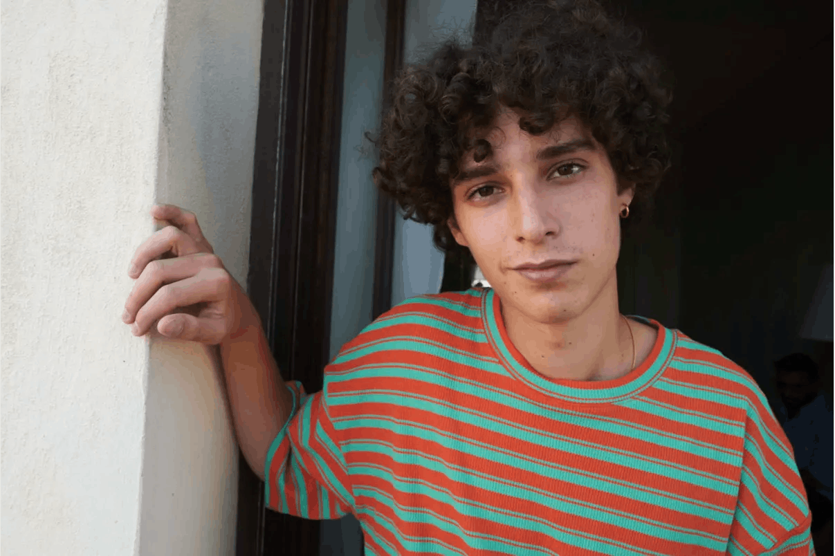 Filippo Scotti wears an orange and turquoise striped shirt as he leans against a doorway and smiles at the camera.