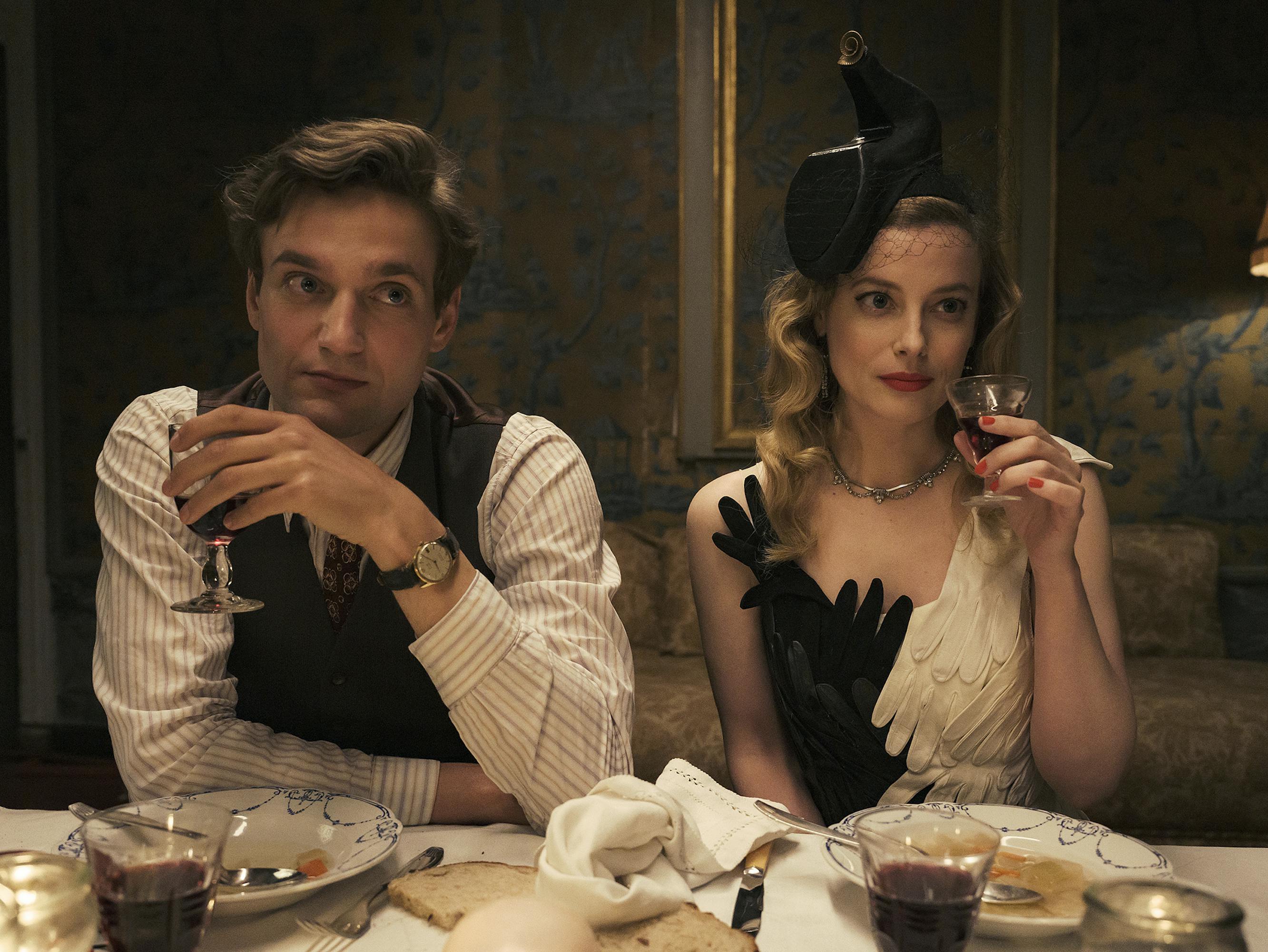 Albert Hirschman (Lucas Englander) and Mary Jayne Gold (Gillian Jacobs) sit at a table sipping red wine in black-and-white outfits. 