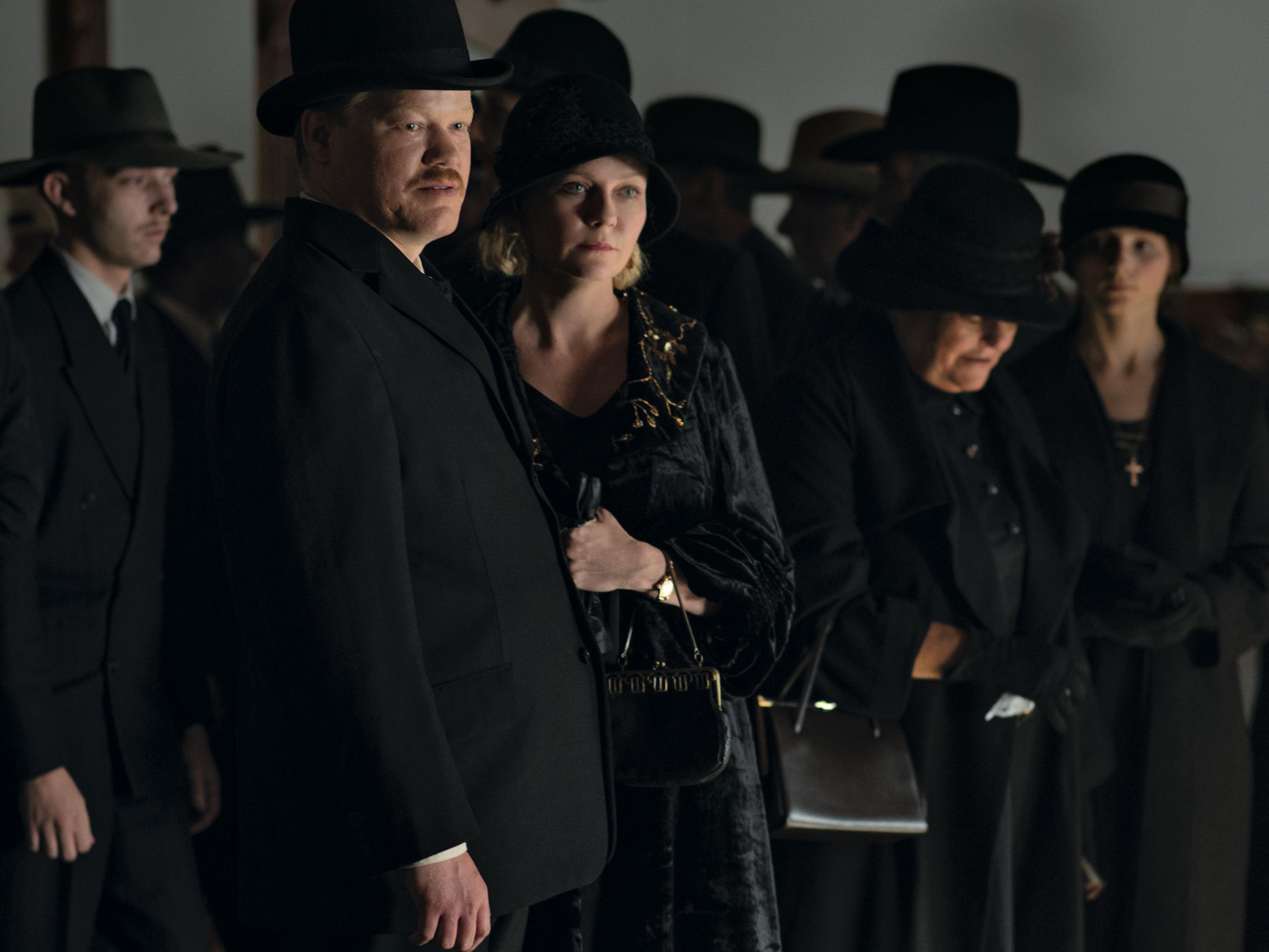 Jesse Plemons and Kirsten Dunst stand with a group of people wearing black outfits and black hats looking somber.