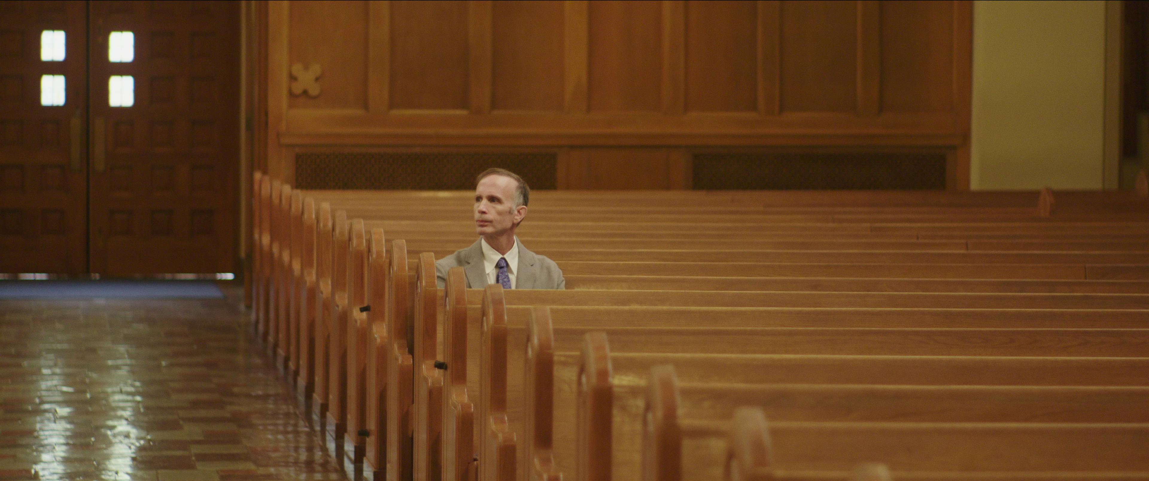 A man sits in otherwise empty church pews. He wears a light grey suit and purple tie. 