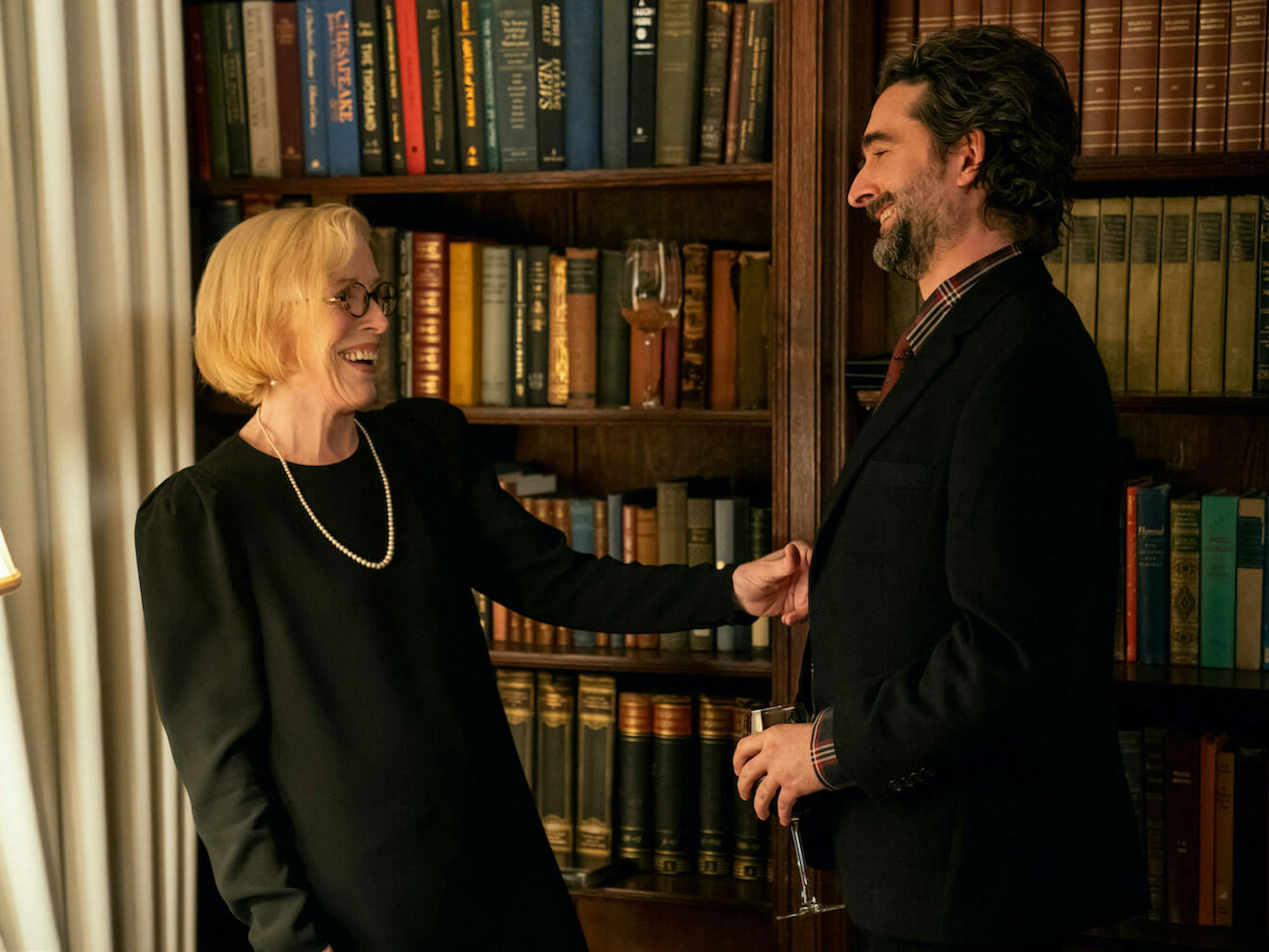 Professor Joan Hambling (Holland Taylor) and Bill Dobson (Jay Duplass) laugh together in all black garb. Behind them lay rows of old books. Duplass holds a glass of wine.